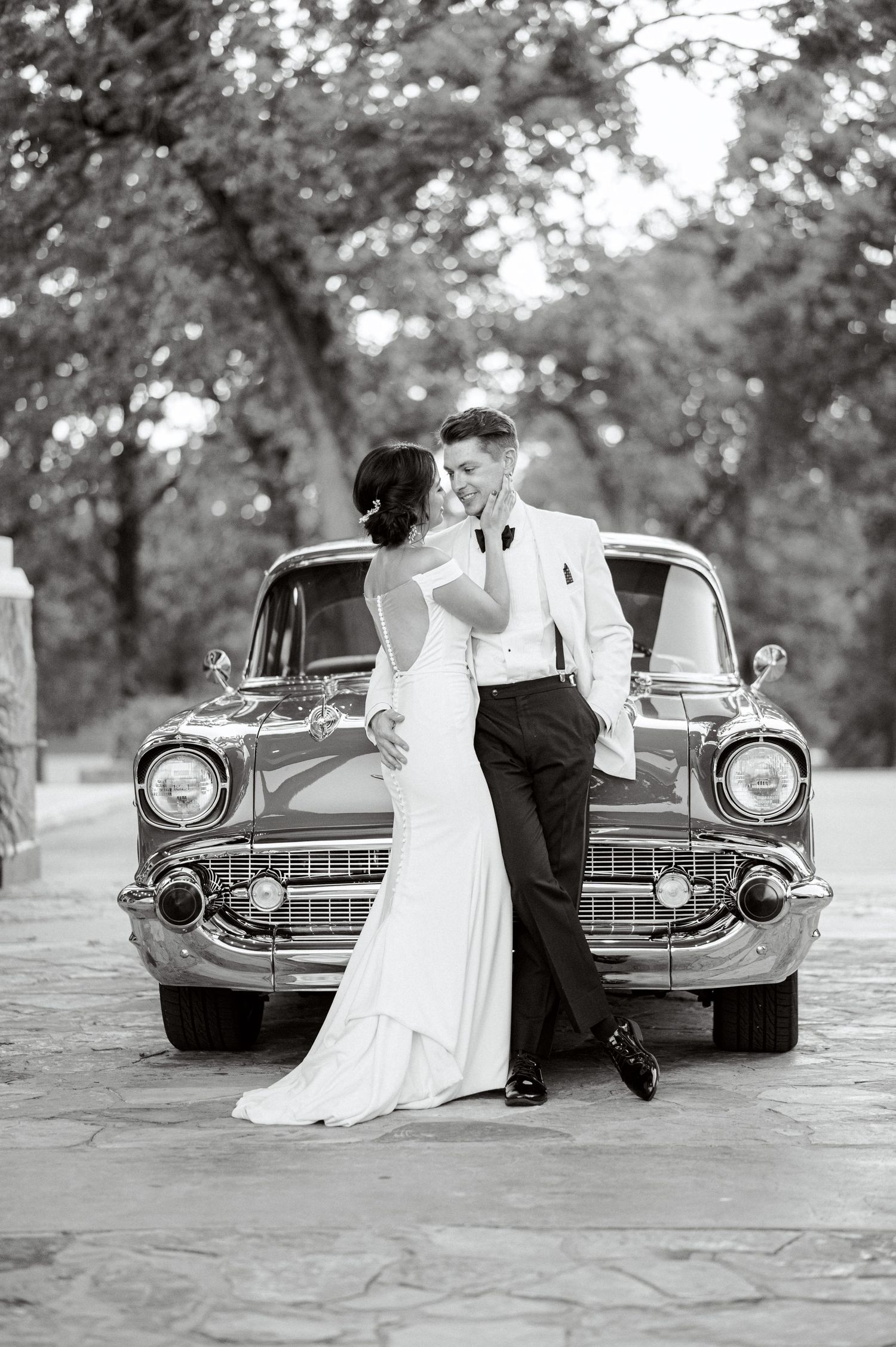 A couple in wedding attire poses romantically in front of a vintage Chevrolet car in black and white photography.