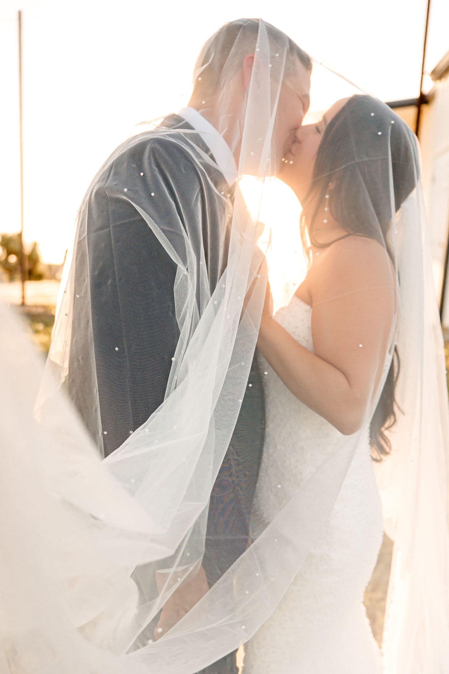 A romantic wedding couple embraces under a flowing white veil during sunset creating a dreamy silhouette effect.
