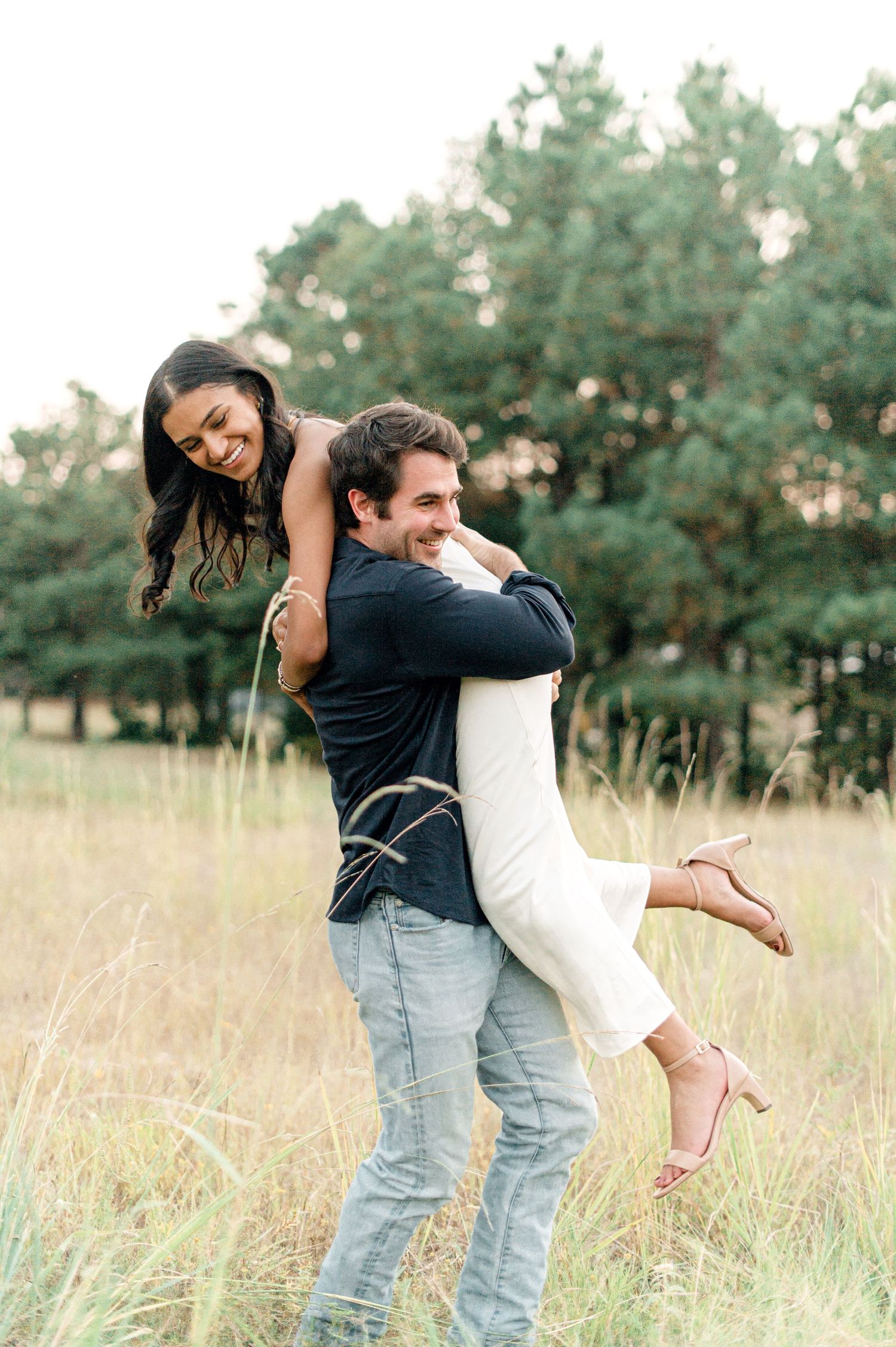 A couple in jeans and white shirt embrace playfully in a golden grass field during sunset.