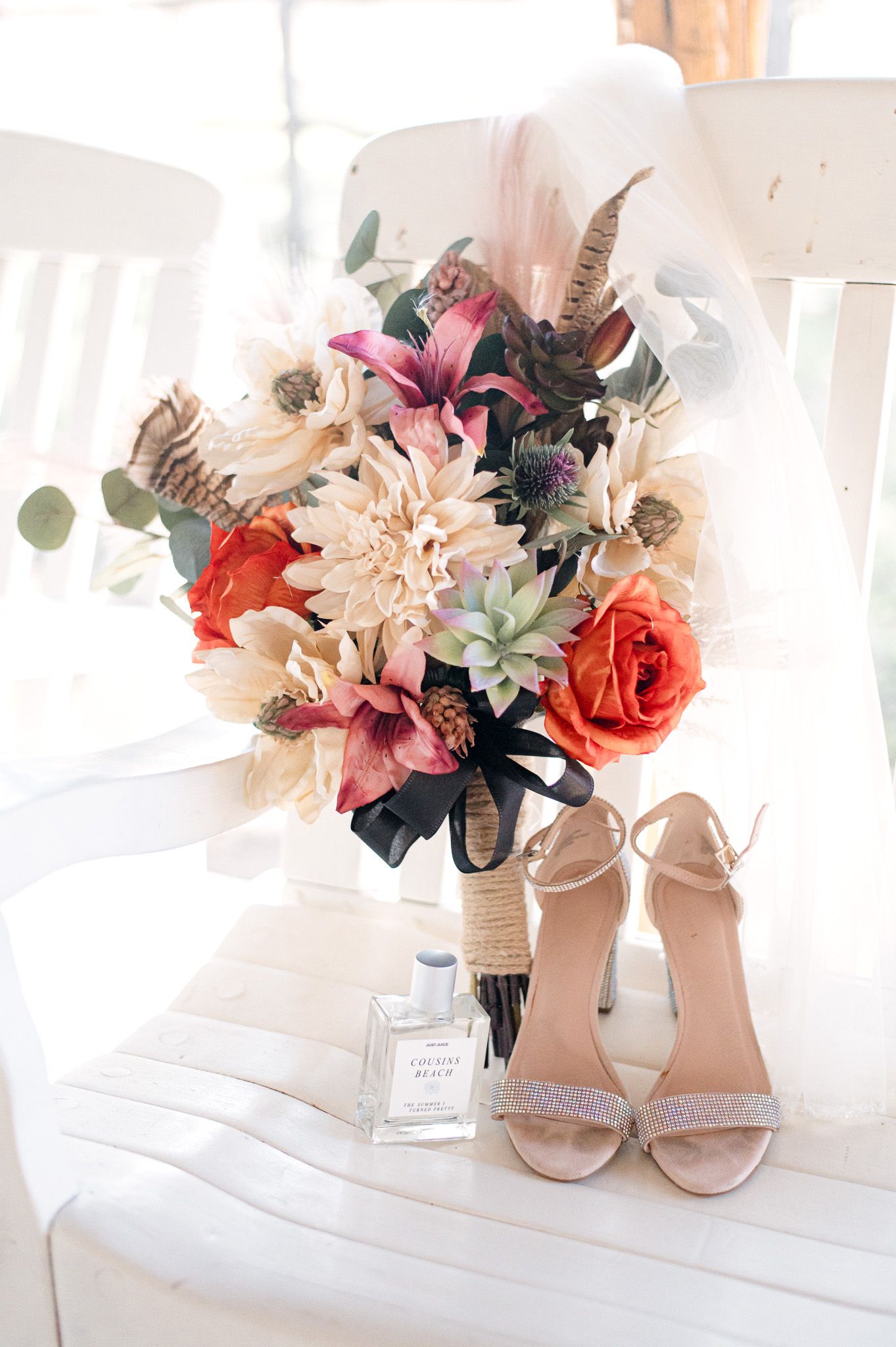 Wedding details featuring a colorful bridal bouquet with roses and feathers, perfume bottle, and nude strappy heels on white chair.