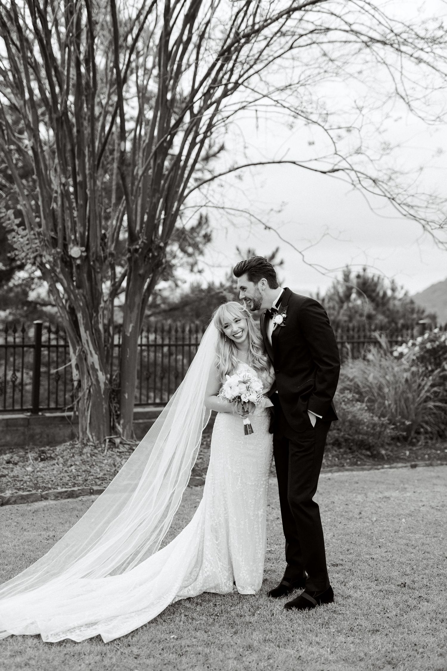 A romantic black and white wedding photo showing a couple in formal attire with a long flowing veil outdoors.  Wedding Photographer tyler tx