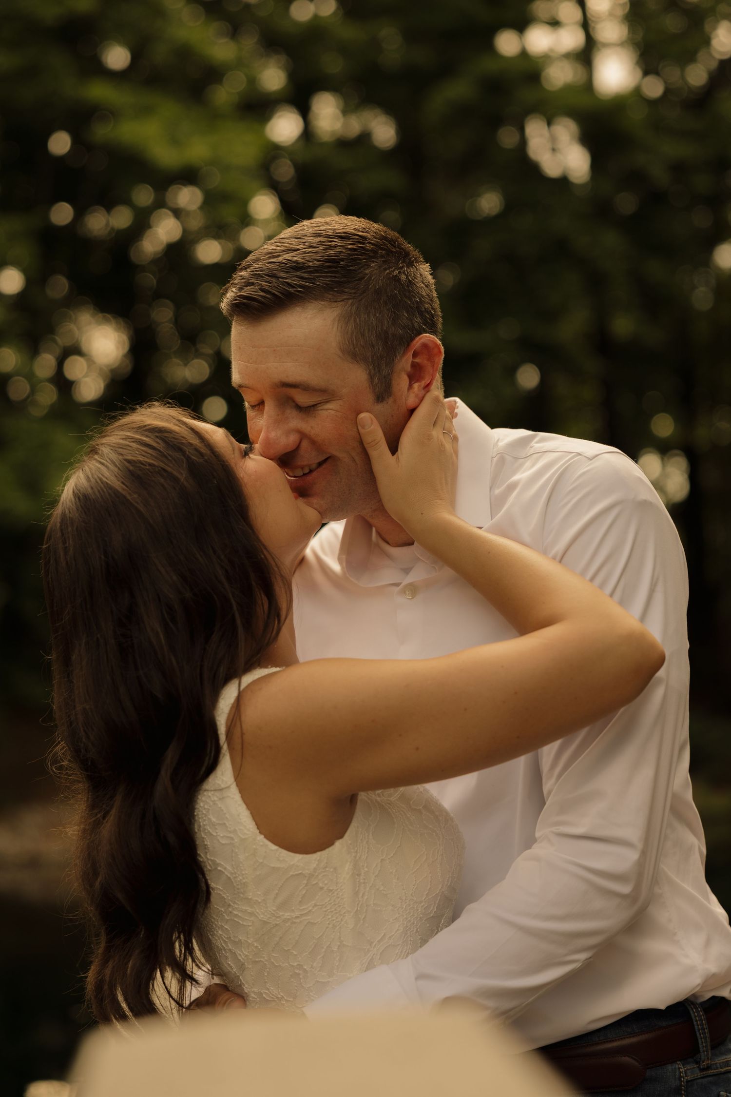 Couple sharing a romantic kiss outdoors, woman in white dress cradling man's face, surrounded by lush green trees.