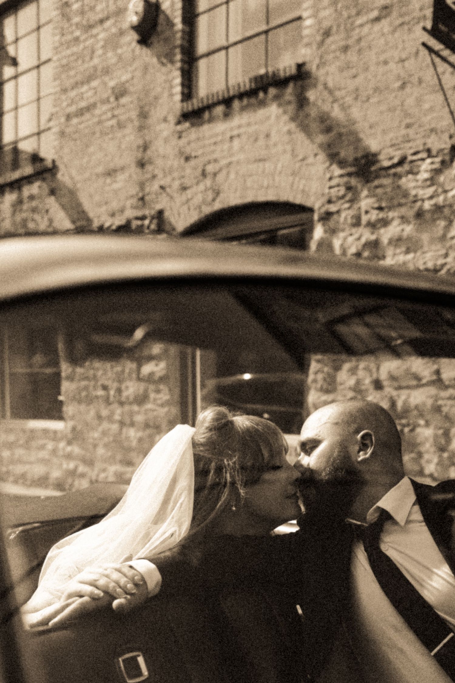Bride and groom sharing a romantic kiss inside a vintage car, with a rustic brick building in the background, sepia tone.