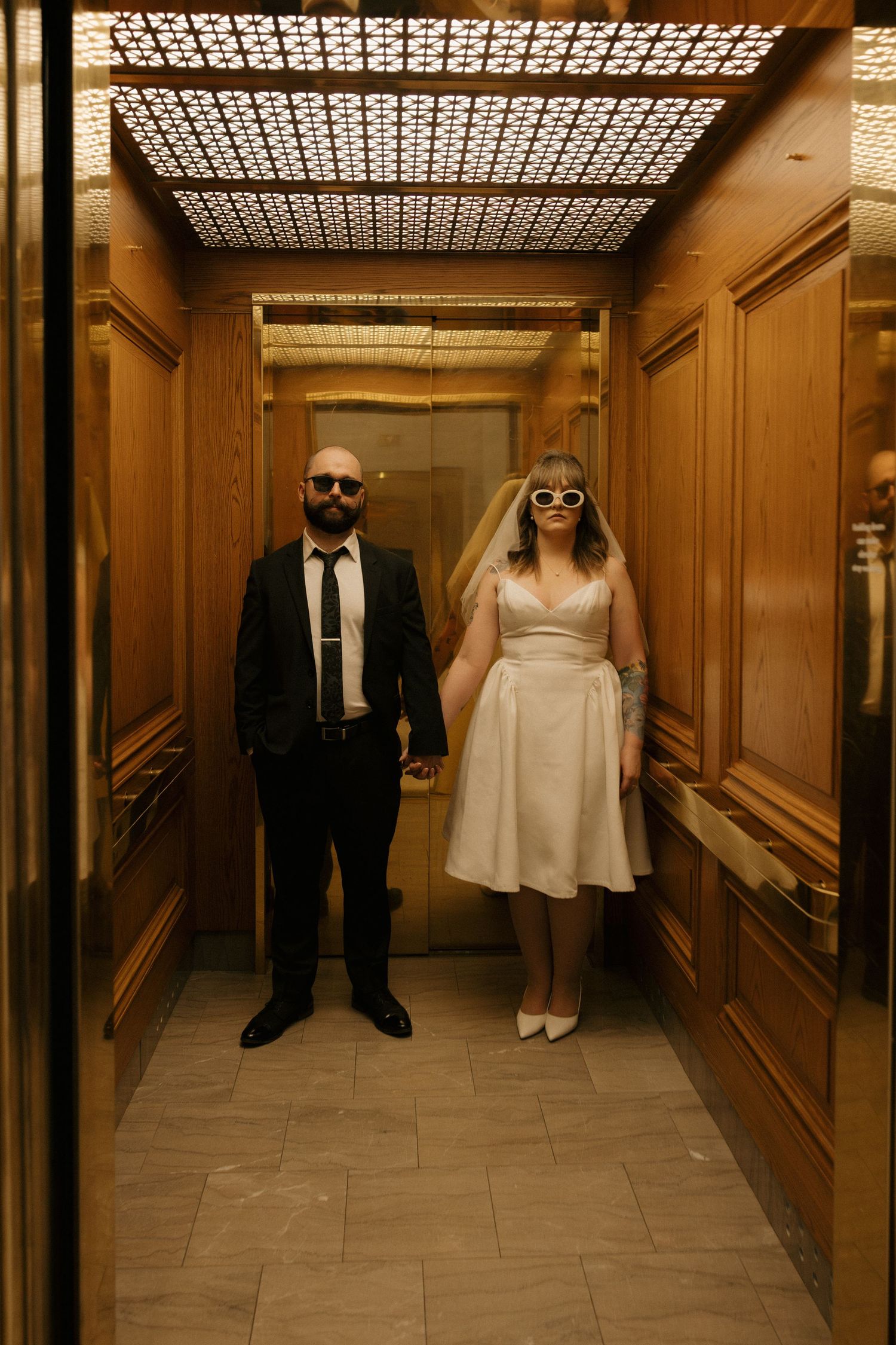 Bride and groom in sunglasses pose together in an elegant wood-paneled elevator on their wedding day.
