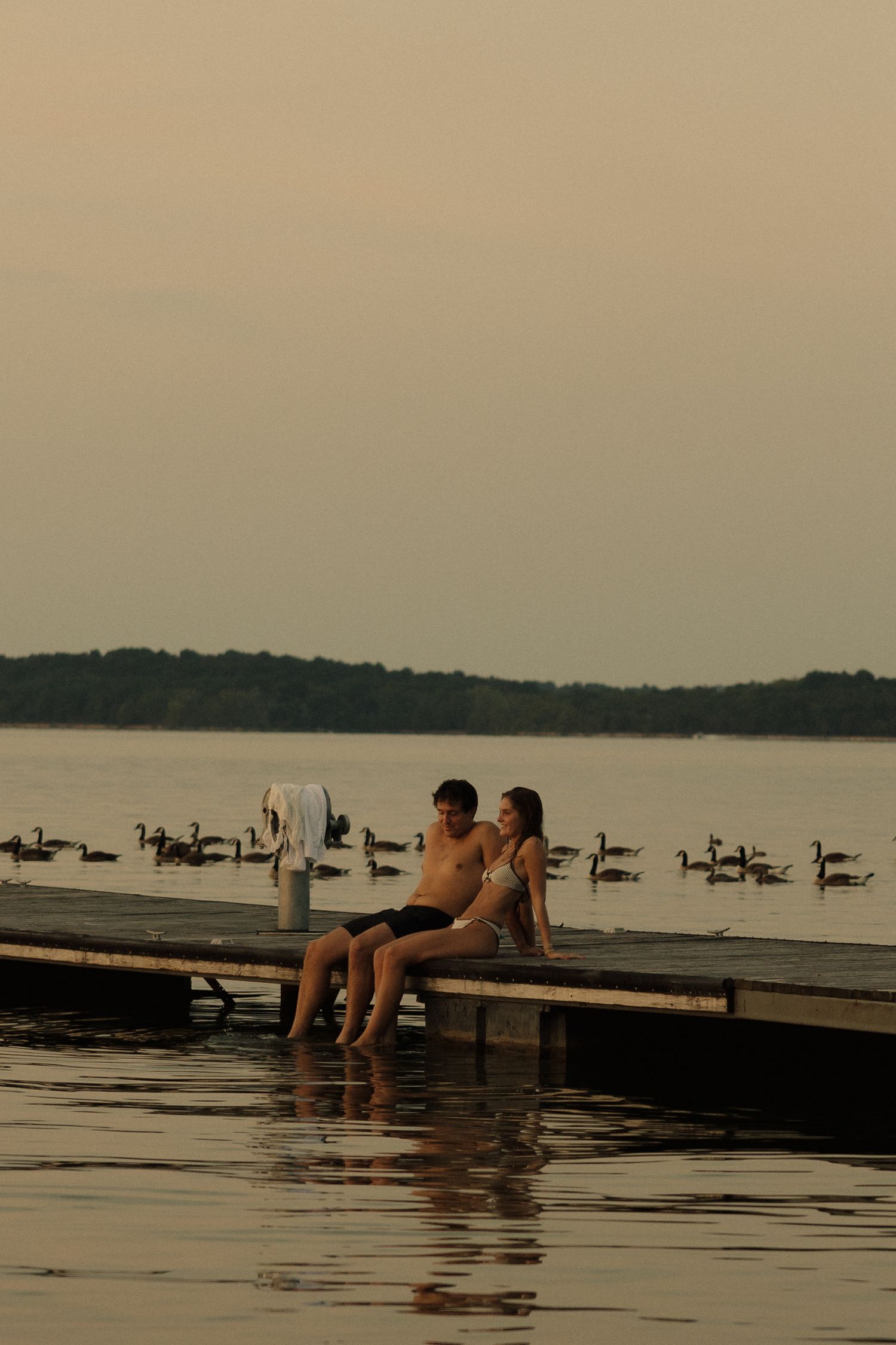 Couple sitting on a wooden dock at sunset with geese swimming in the calm lake behind them.