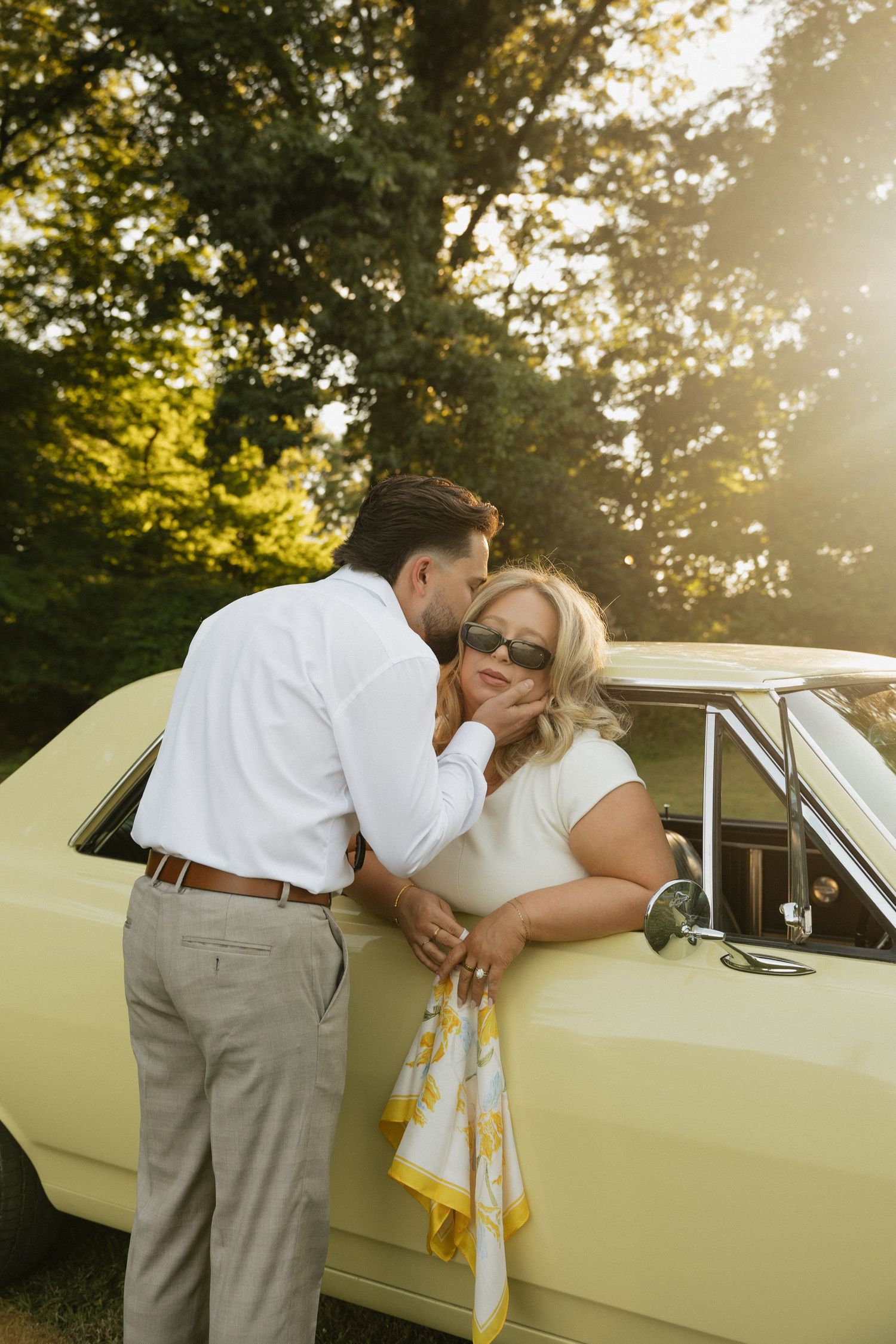 Couple sharing a romantic moment leaning against a vintage yellow car surrounded by lush green trees in golden sunlight.