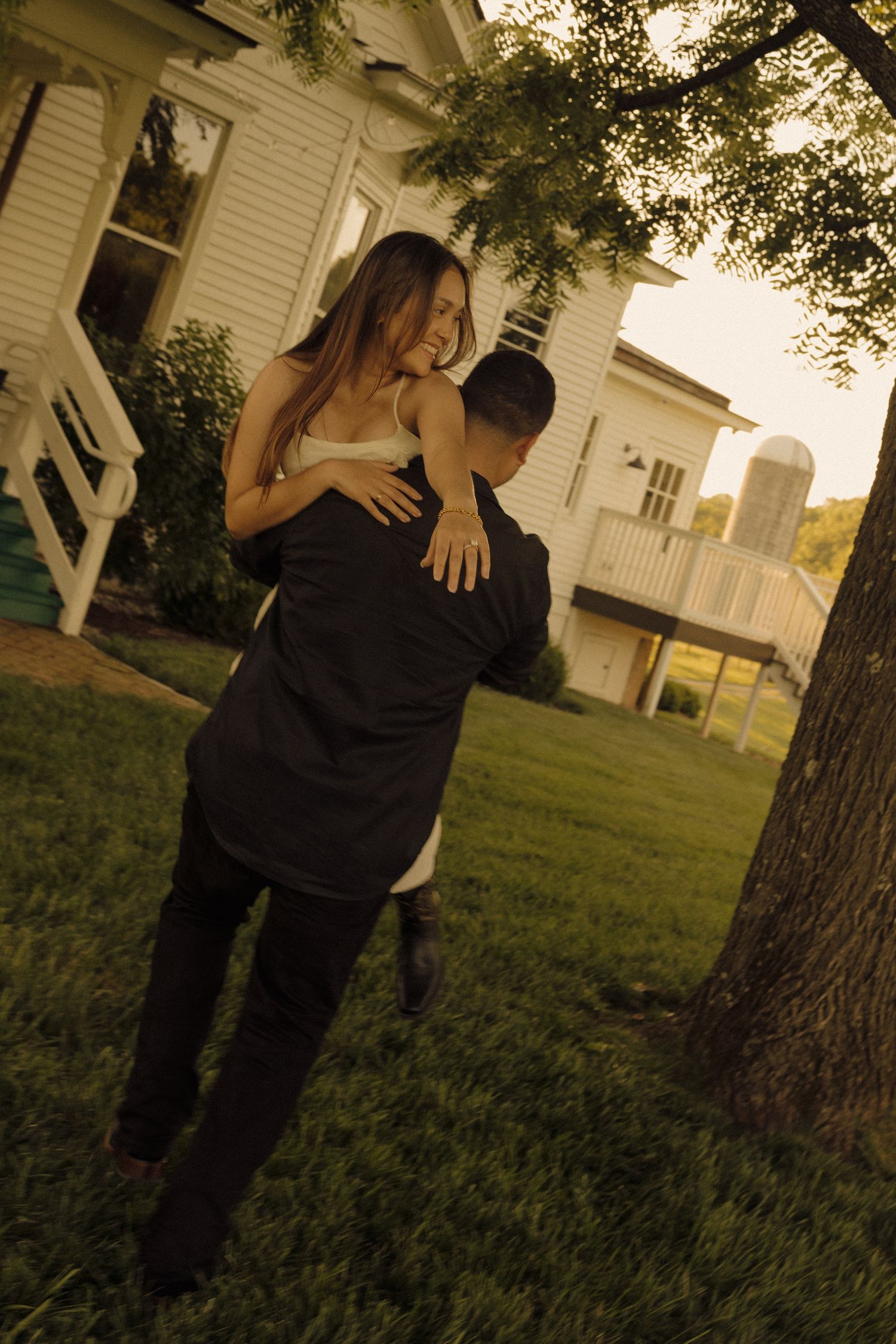A couple embracing and laughing in a green yard outside a white farmhouse at golden hour.