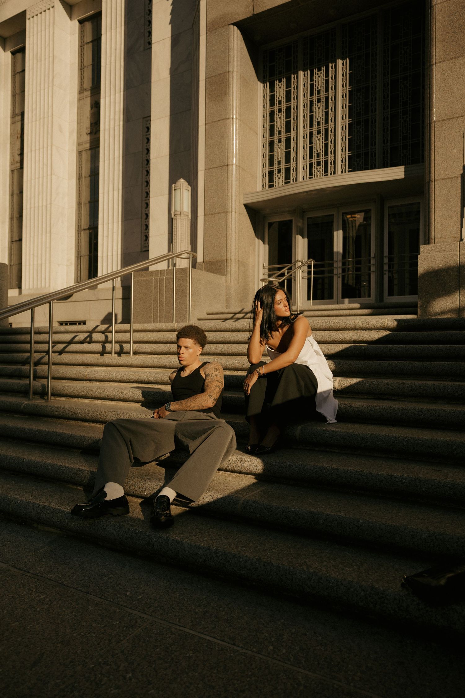 Two people sit on stone steps of a grand building in warm golden light, one with tattoos, both in casual urban outfits.