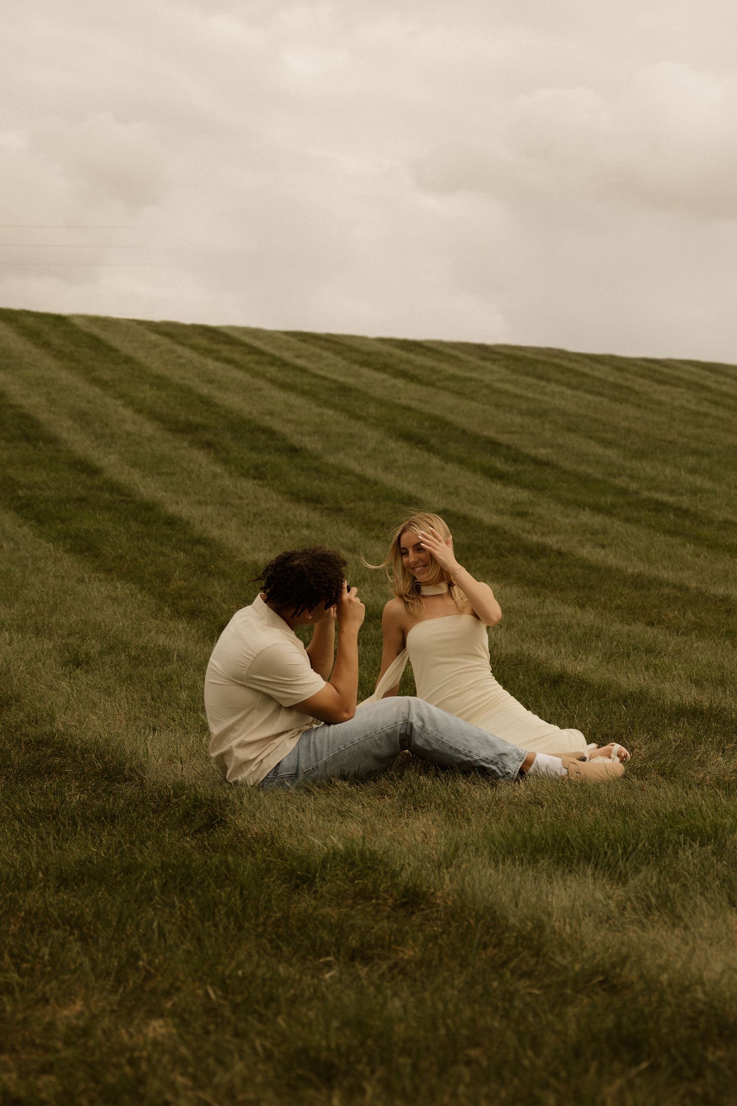 A couple sits on a lush green hillside, man photographing woman as she covers her eyes, under a cloudy sky.