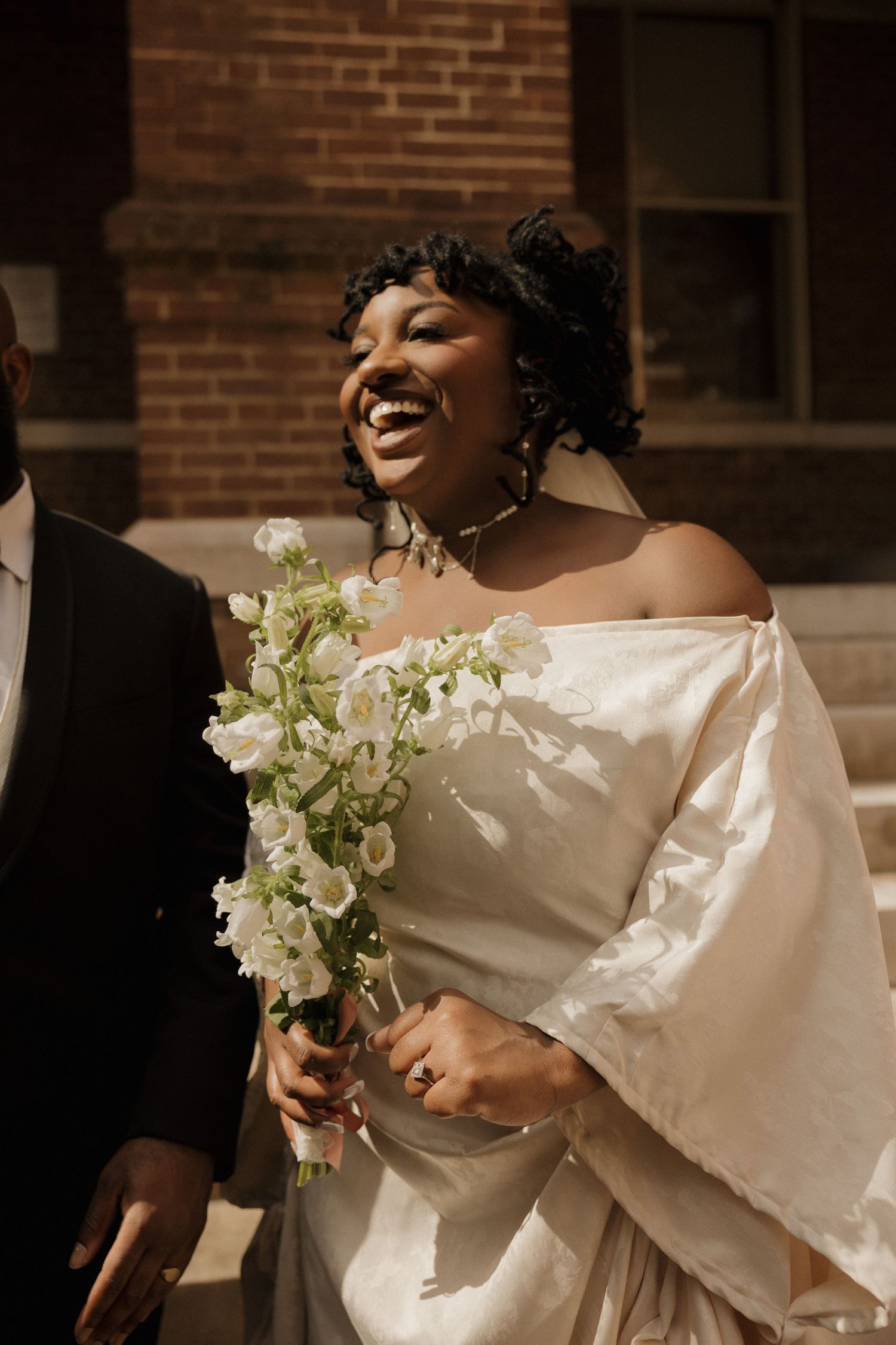 A joyful bride in an off-shoulder wedding dress holds a white and green bouquet against a brick wall backdrop.