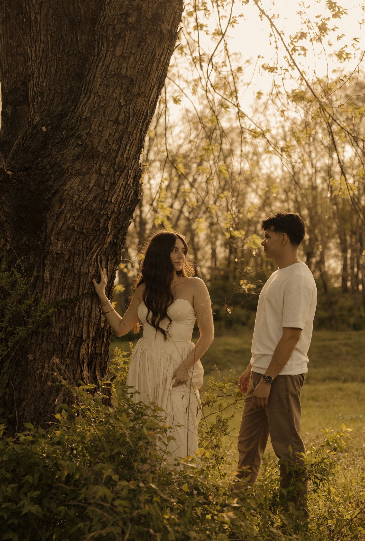 Couple sharing a romantic moment by a large tree in golden hour sunlight amid lush greenery.