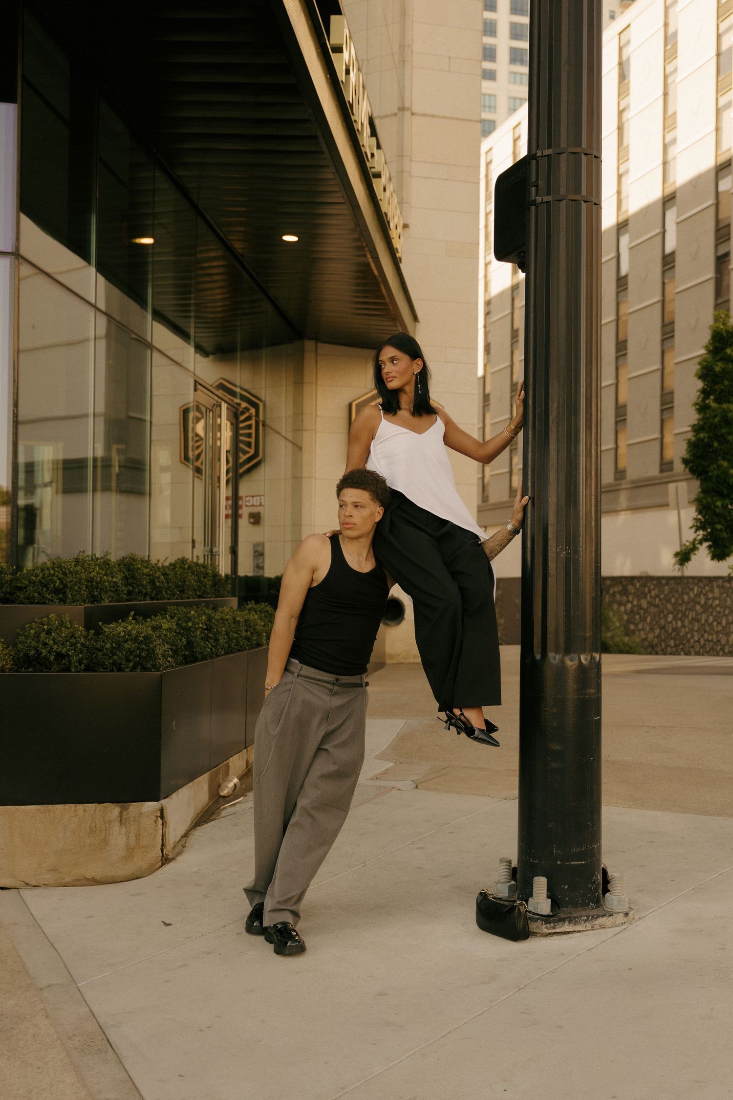 Two stylish people pose on an urban sidewalk, one leaning on a light pole, wearing monochrome outfits in a city setting.