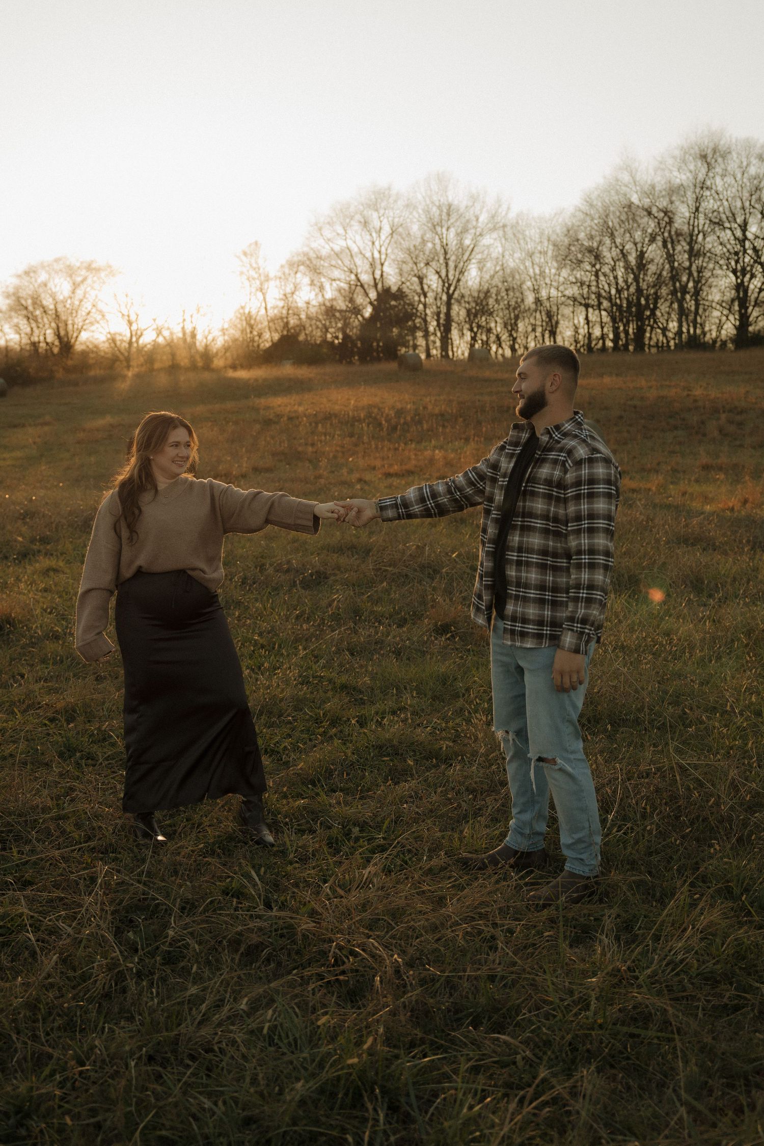 A couple holds hands and dances together in a golden field at sunset during an outdoor photoshoot.