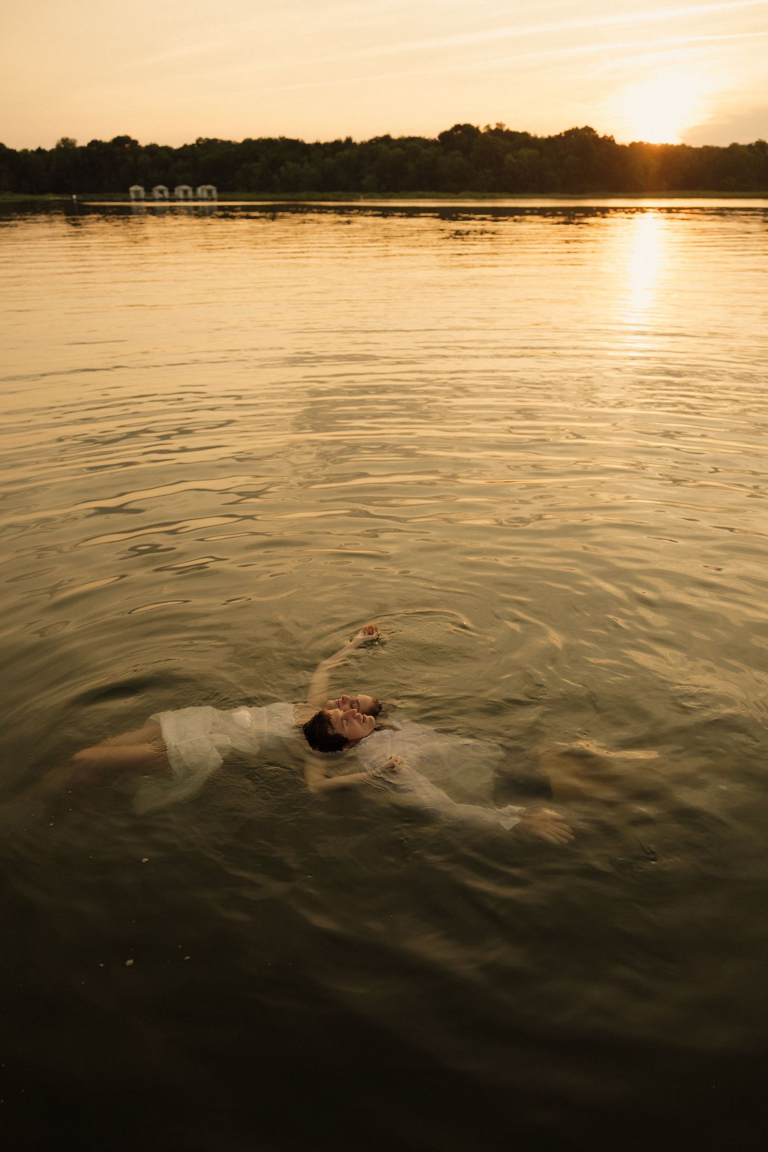 Person floating on their back in a calm lake wearing a white dress during a golden sunset with trees in the background.
