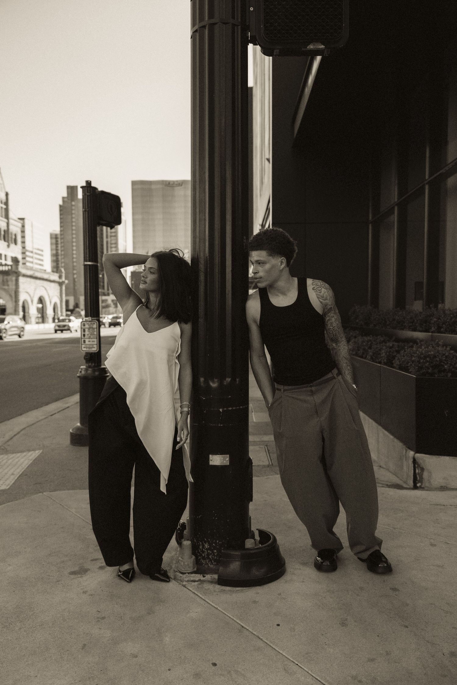Black and white photo of a man and woman leaning against a street pole on an urban sidewalk with city buildings behind.