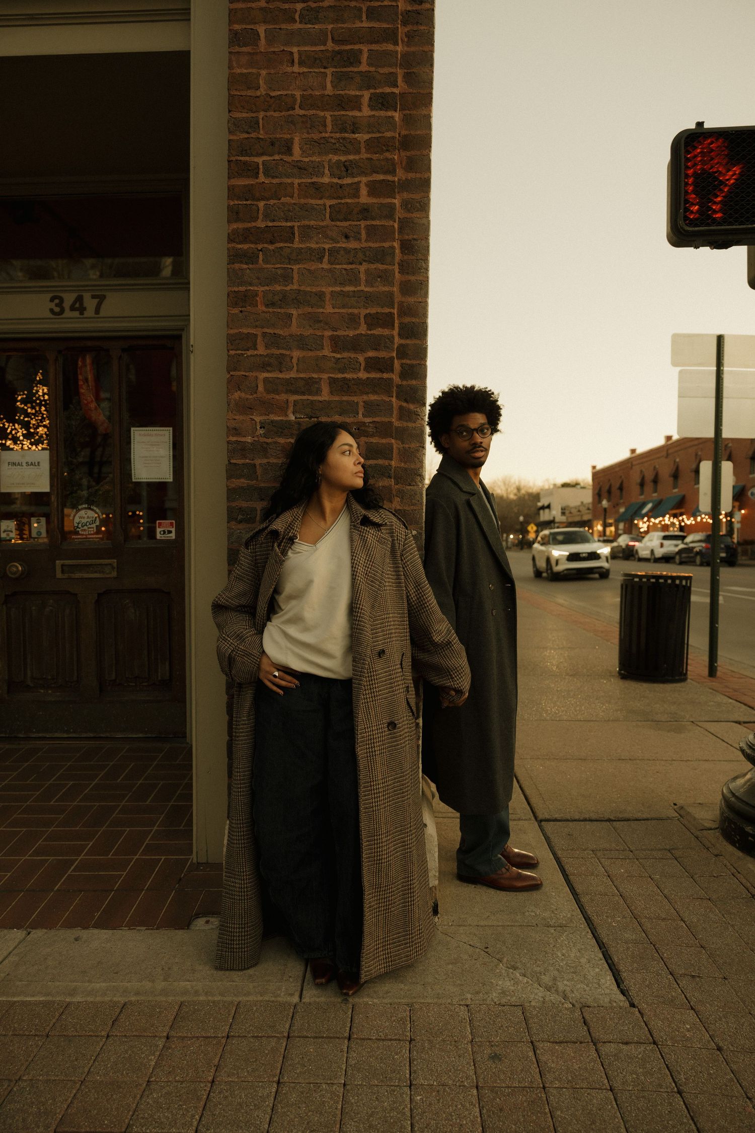 Two people in long coats stand on a city sidewalk at dusk near a brick building numbered 347.