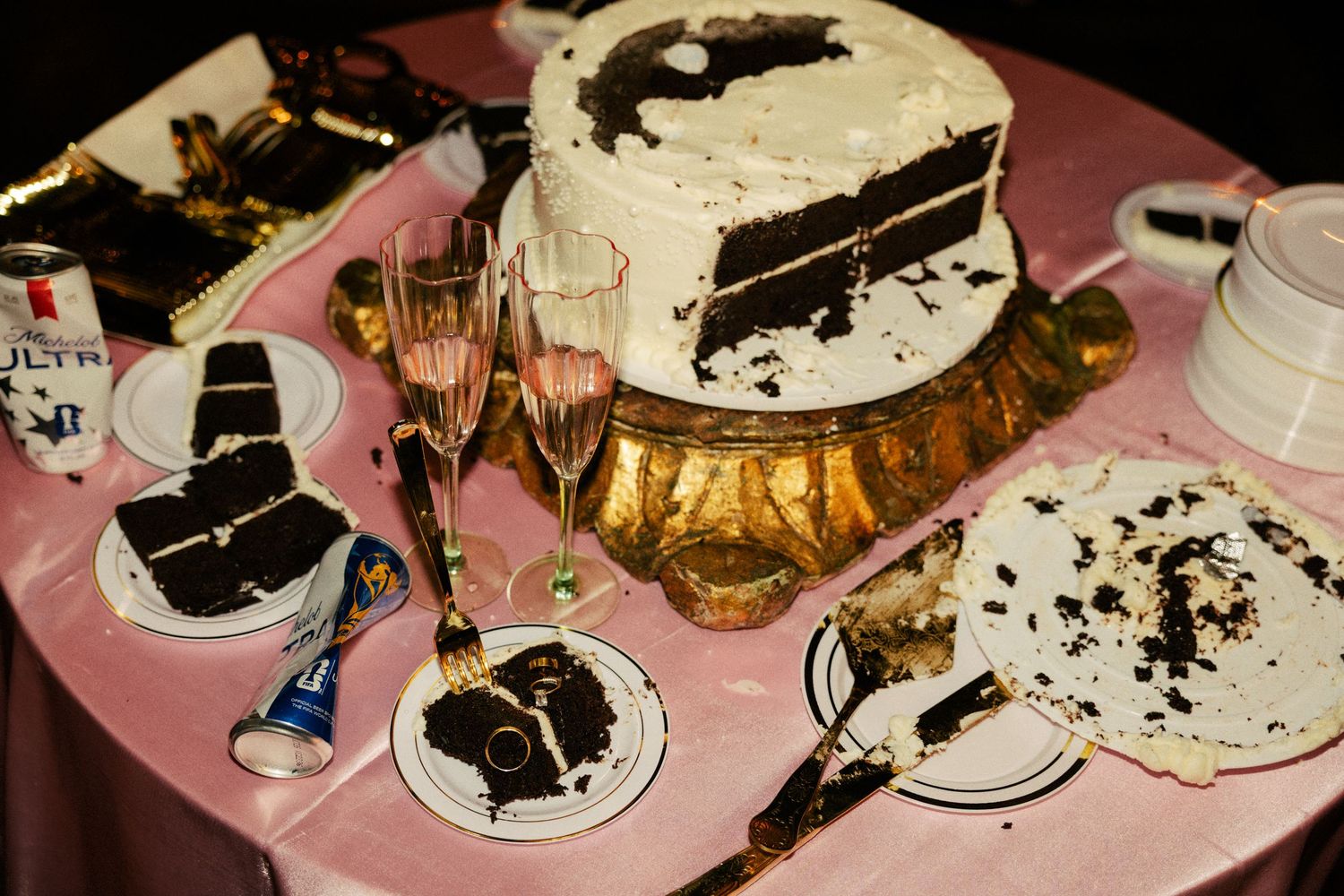 Elegant chocolate cake with white frosting on gold stand, champagne flutes, and served slices on pink tablecloth.