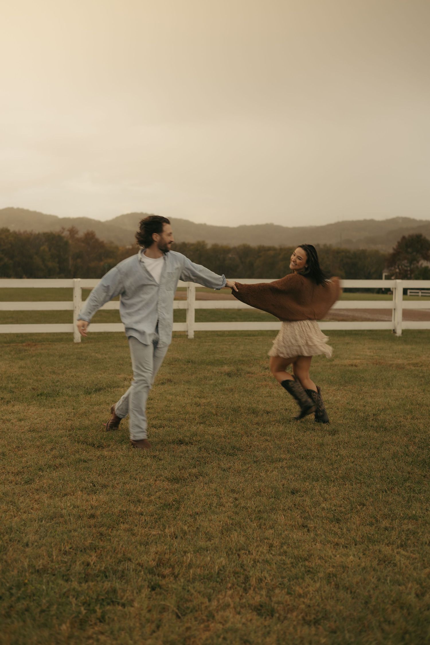 Two people dance together playfully in a grassy field with a white fence and rolling hills in the background at sunset.