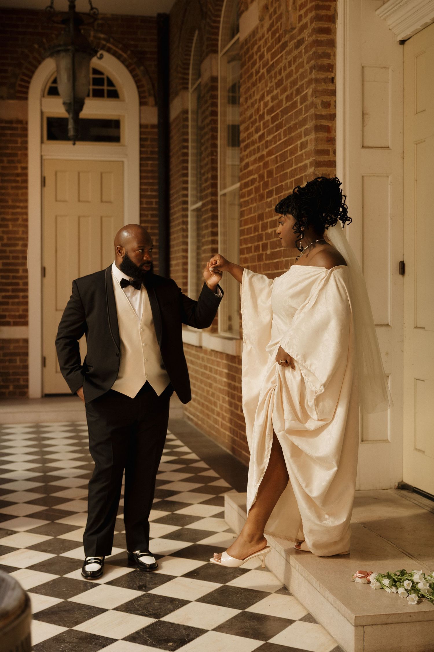 Wedding couple poses playfully on black and white checkered floor in elegant venue hallway.