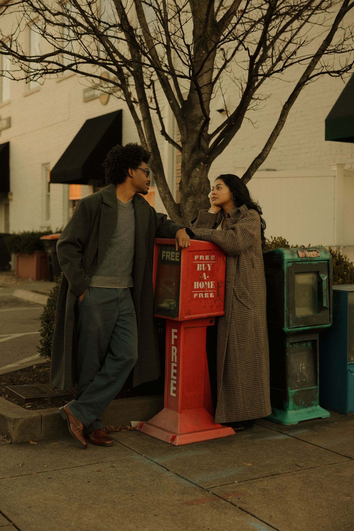 Two young people in long coats chat beside a red free newspaper stand on a city sidewalk near a bare tree.