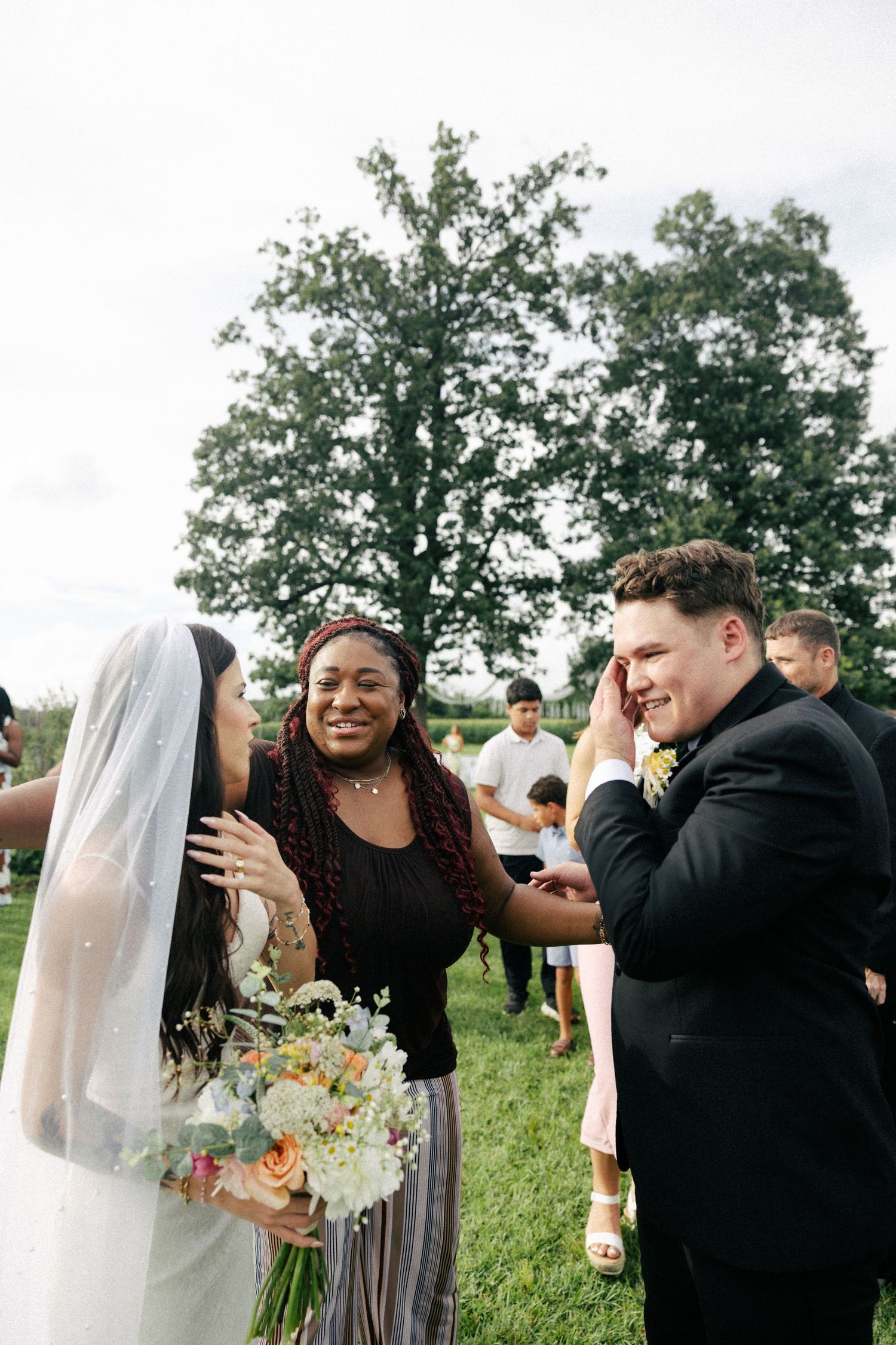 Bride and groom sharing emotional moment with guest at outdoor wedding ceremony, bride holding colorful bouquet.