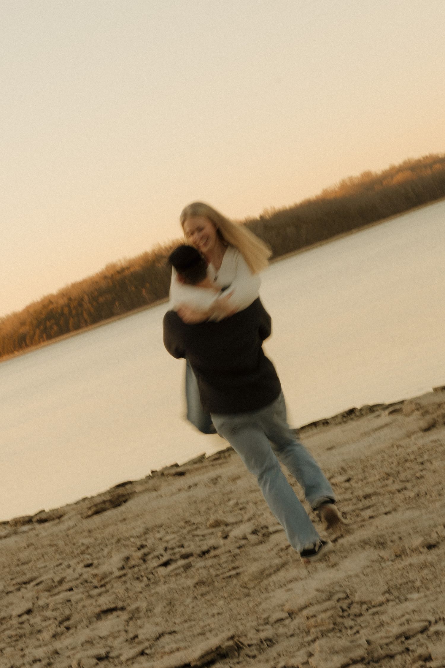 A person in a black jacket and jeans skips rocks on a beach at sunset with a calm lake in the background.