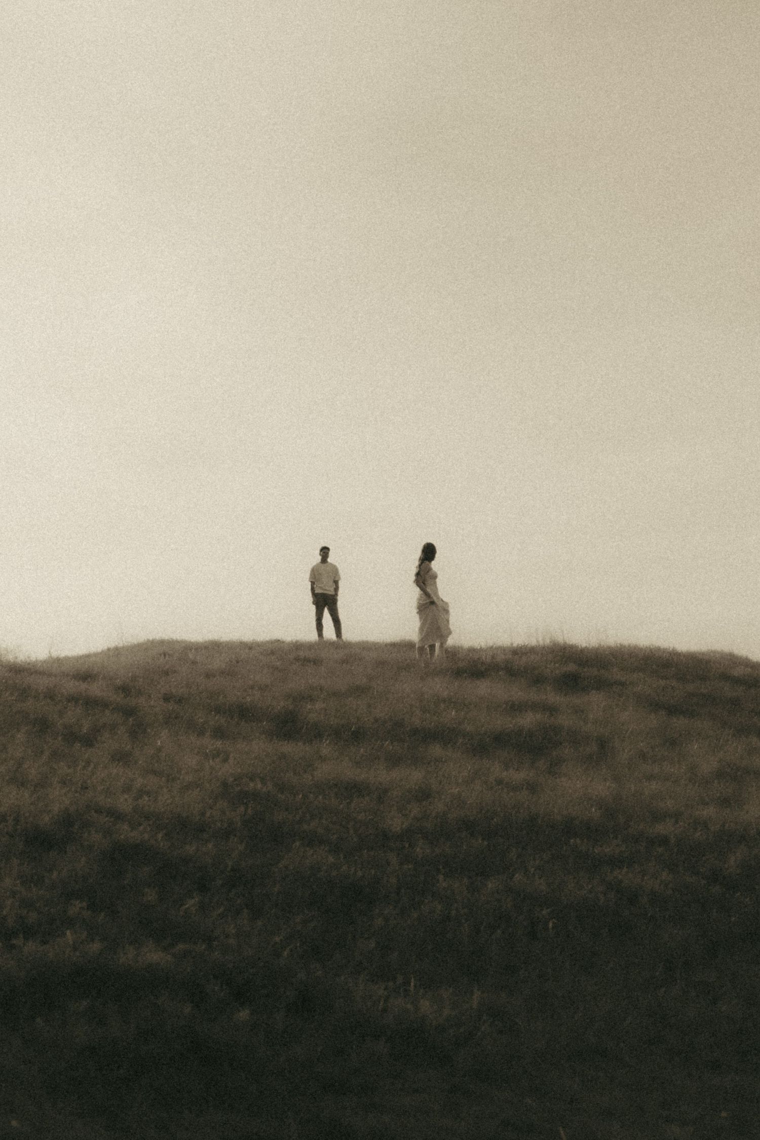 Two figures stand apart on a misty grassy hill in a moody sepia-toned landscape photograph.