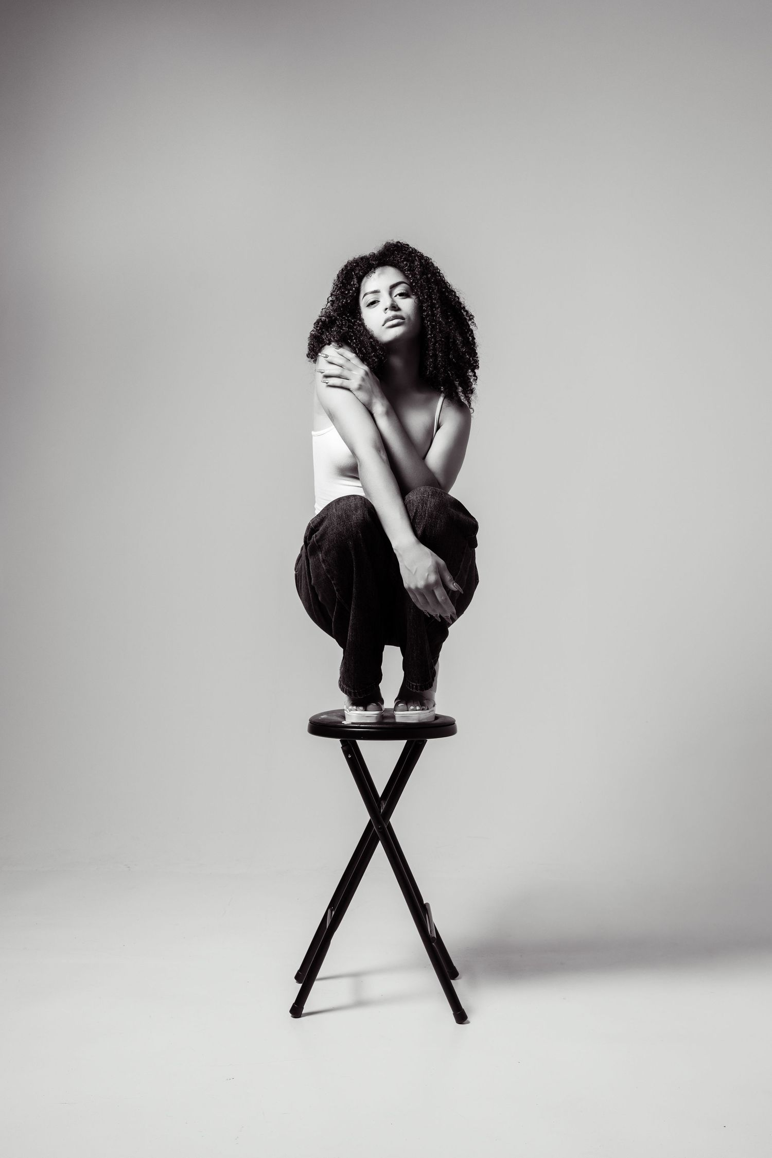 Black and white photo of a woman with curly hair squatting on a stool, posing gracefully in a studio setting.