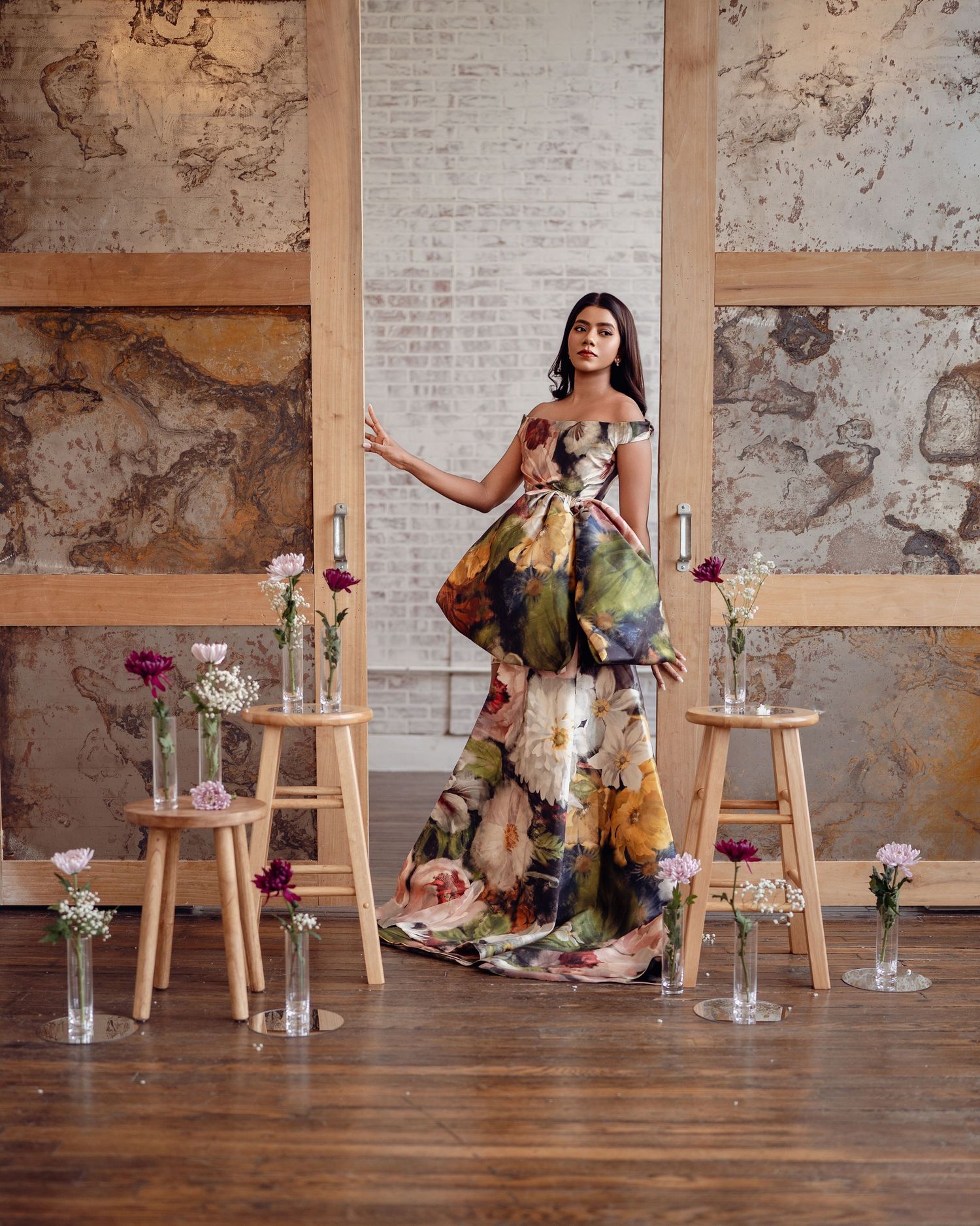 Woman in floral off-shoulder gown poses among bud vases on wooden stools in rustic industrial setting.