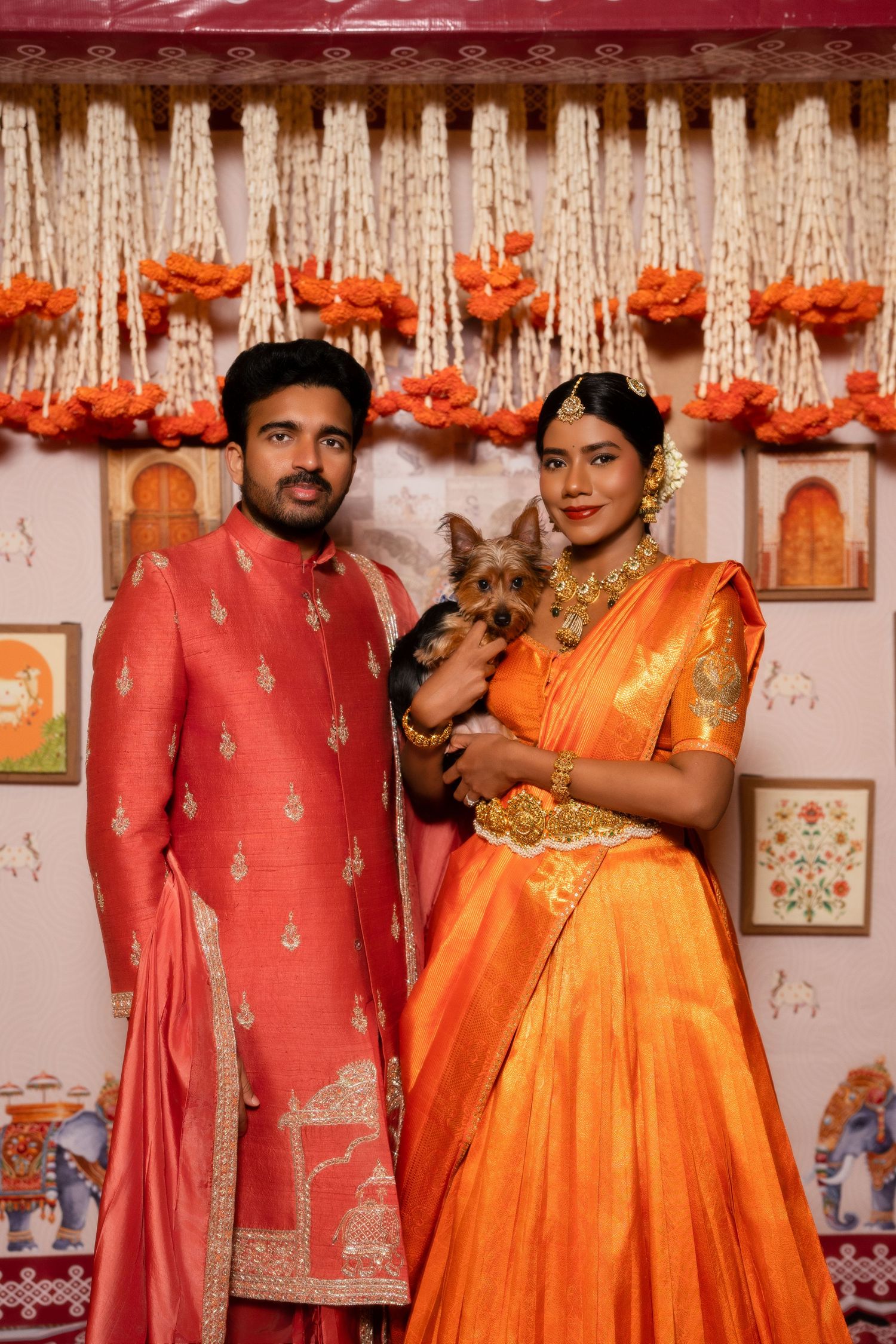 Indian couple in traditional orange and red attire posing with a small dog at a festive celebration.
