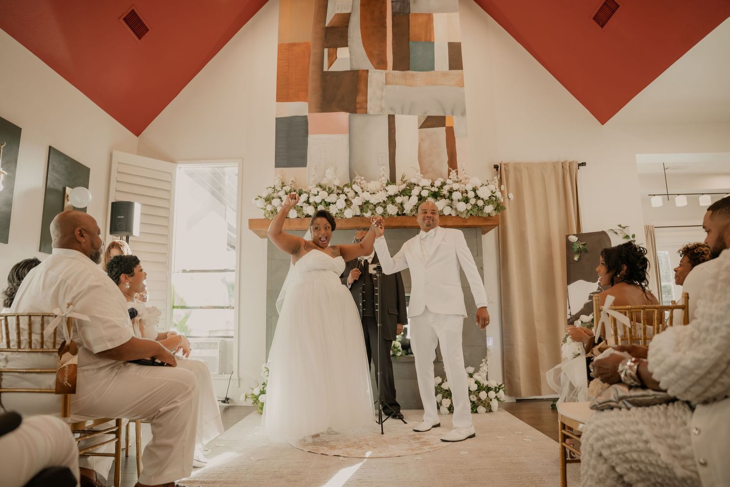 Couple exchanges vows at elegant indoor wedding ceremony with floral arch, guests seated in white chairs, colorful geometric backdrop.