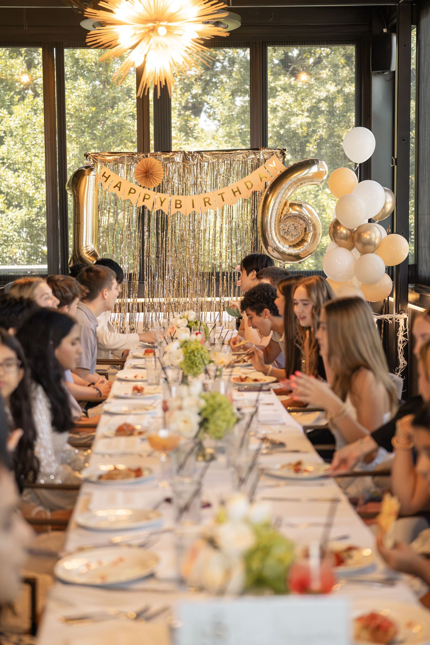 Teens celebrate a 16th birthday party at a long decorated table with gold balloons and Happy Birthday banner.