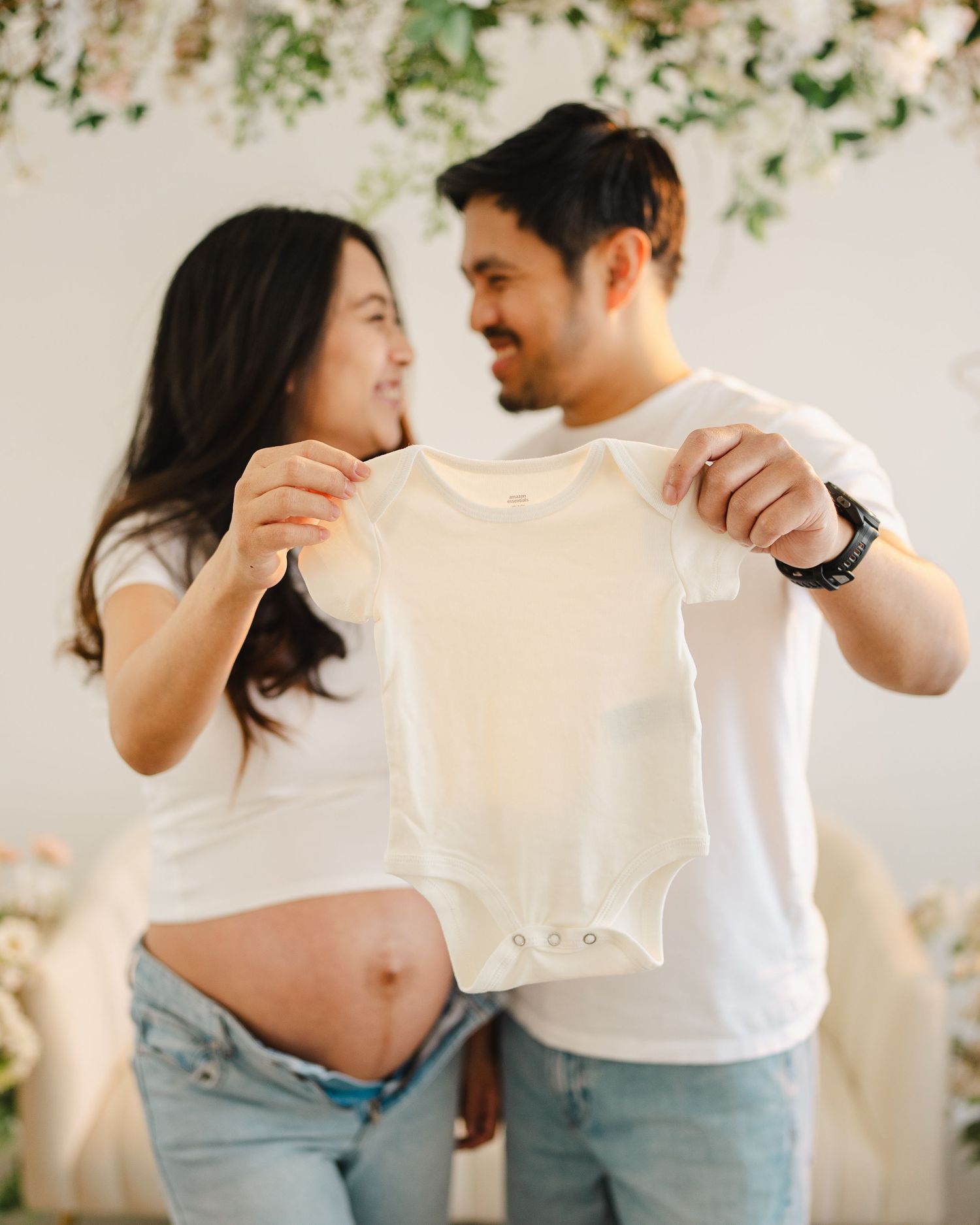 Expecting couple holding white baby onesie together, smiling at each other during maternity photo shoot with floral backdrop.