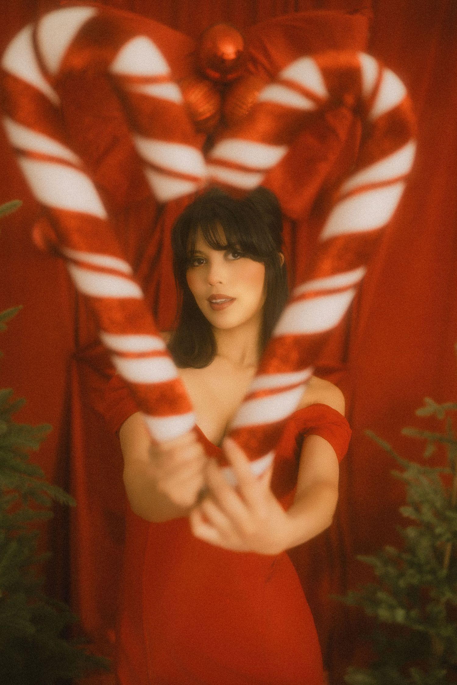 Woman in red off-shoulder dress holds giant candy canes forming a heart shape against a festive red backdrop.
