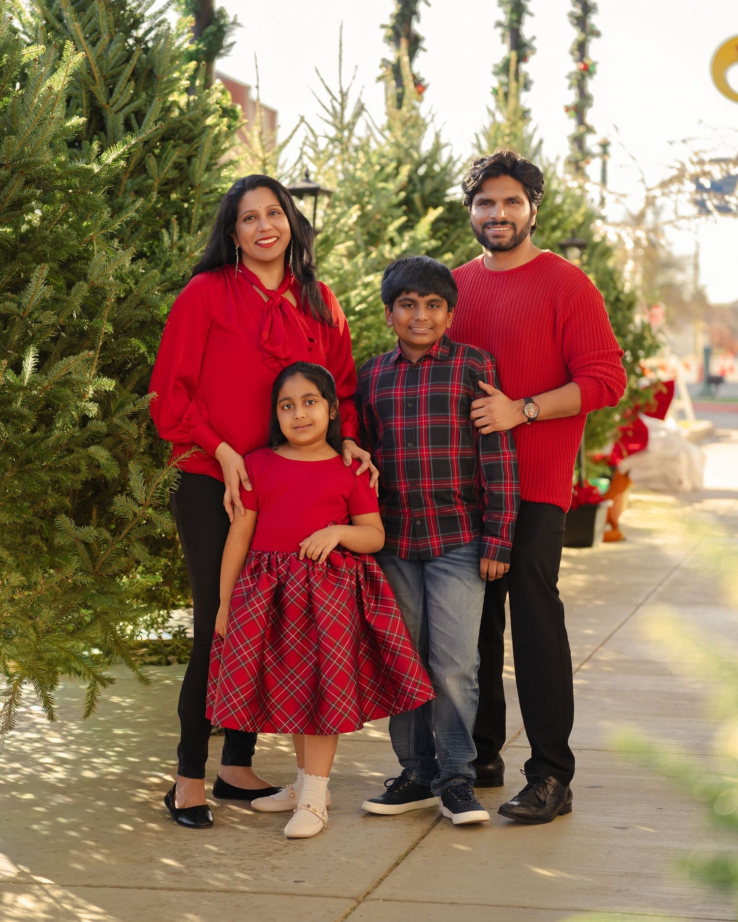 Family of four in red outfits posing together at a Christmas tree lot during the holiday season.