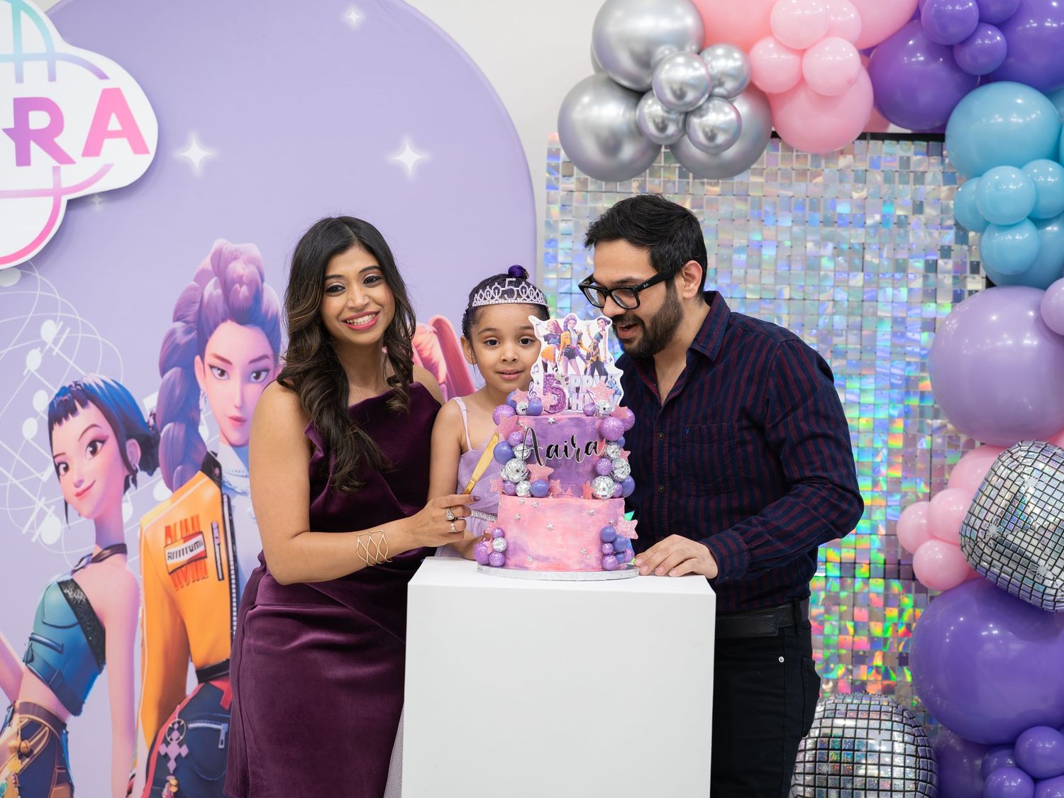 A festive birthday party celebration with pink and purple balloon decorations and people gathered around a cake.