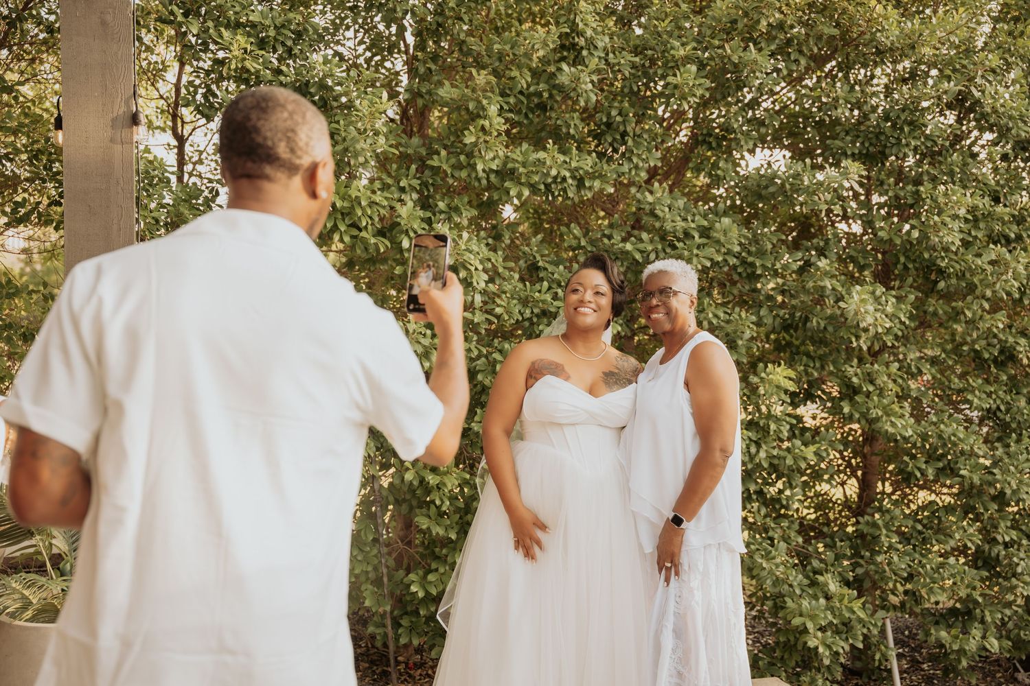 A man photographs two women in white dresses smiling outdoors near green trees at a wedding celebration.