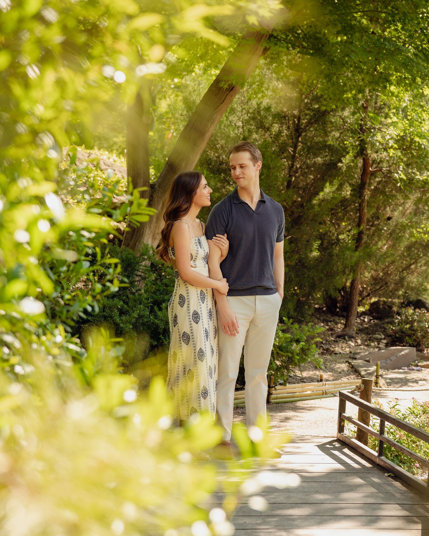Couple standing on a wooden bridge surrounded by lush green trees in a sunlit garden setting.