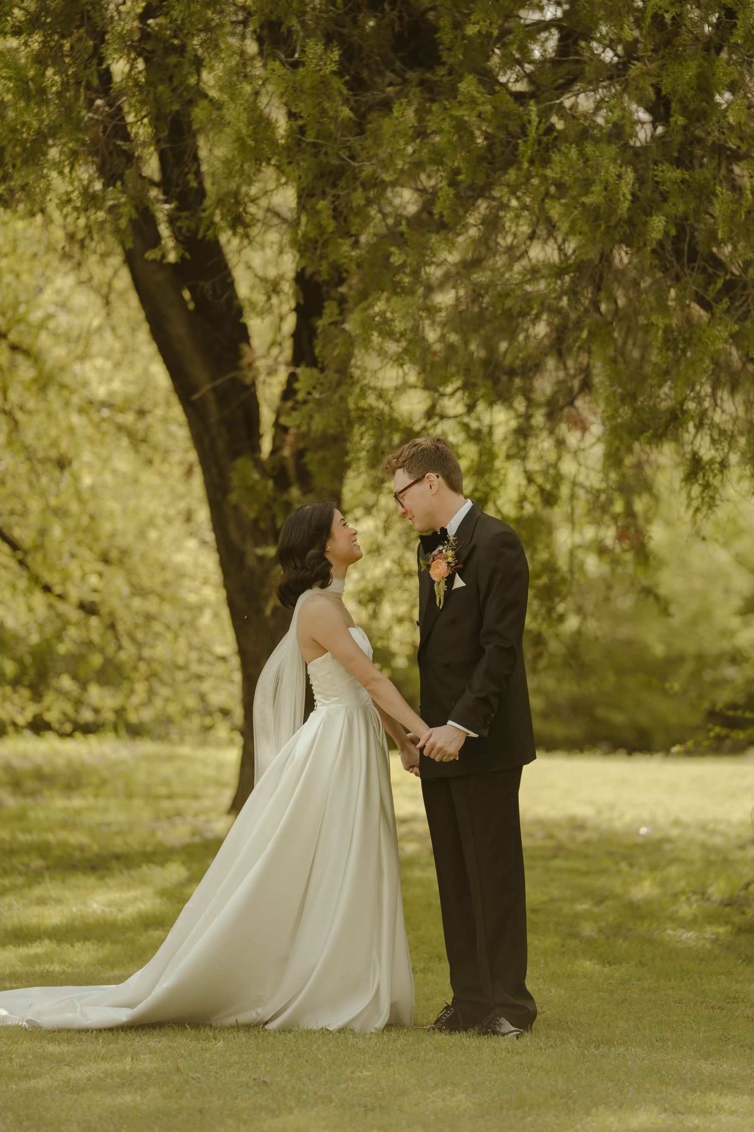 Bride and groom hold hands under a willow tree on green grass, exchanging loving glances in wedding attire.