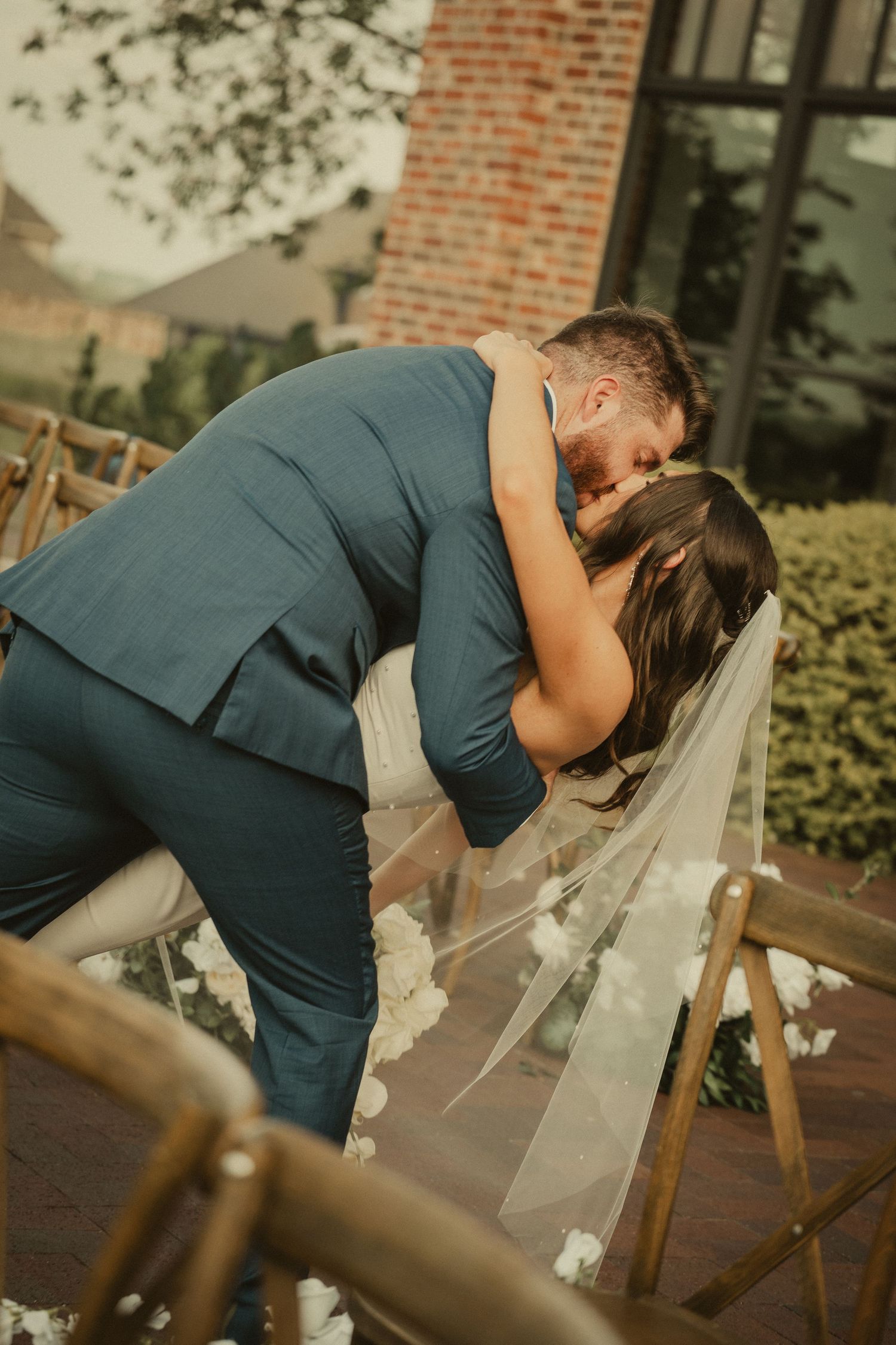 Groom in blue suit dips bride in white veil for romantic kiss at outdoor wedding ceremony.