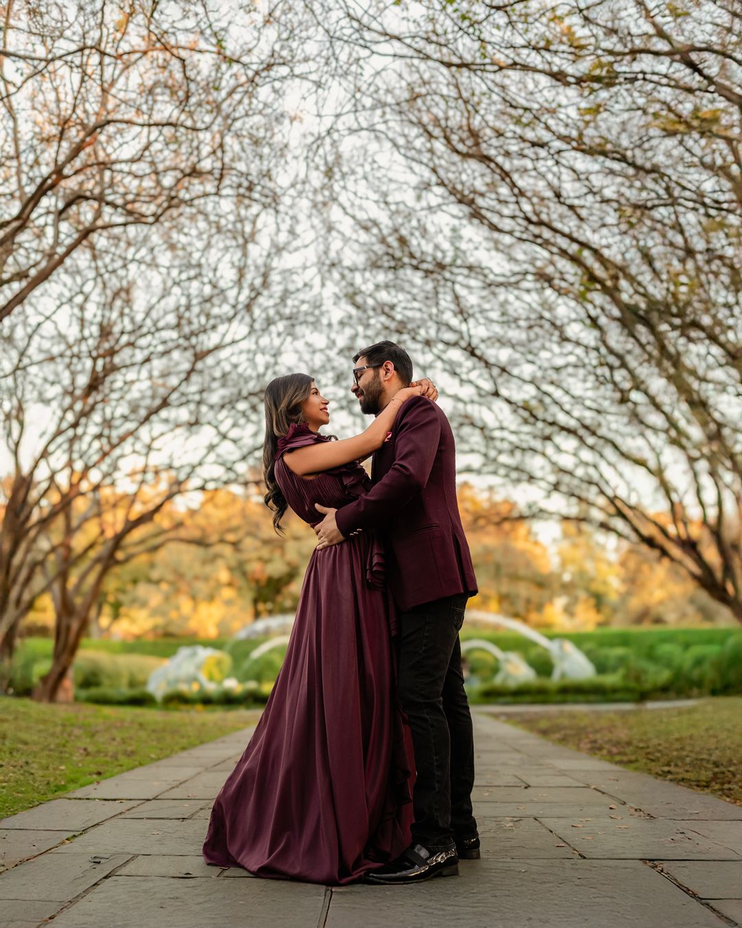Romantic couple in burgundy and black outfits sitting together on a stone pathway surrounded by autumn trees.