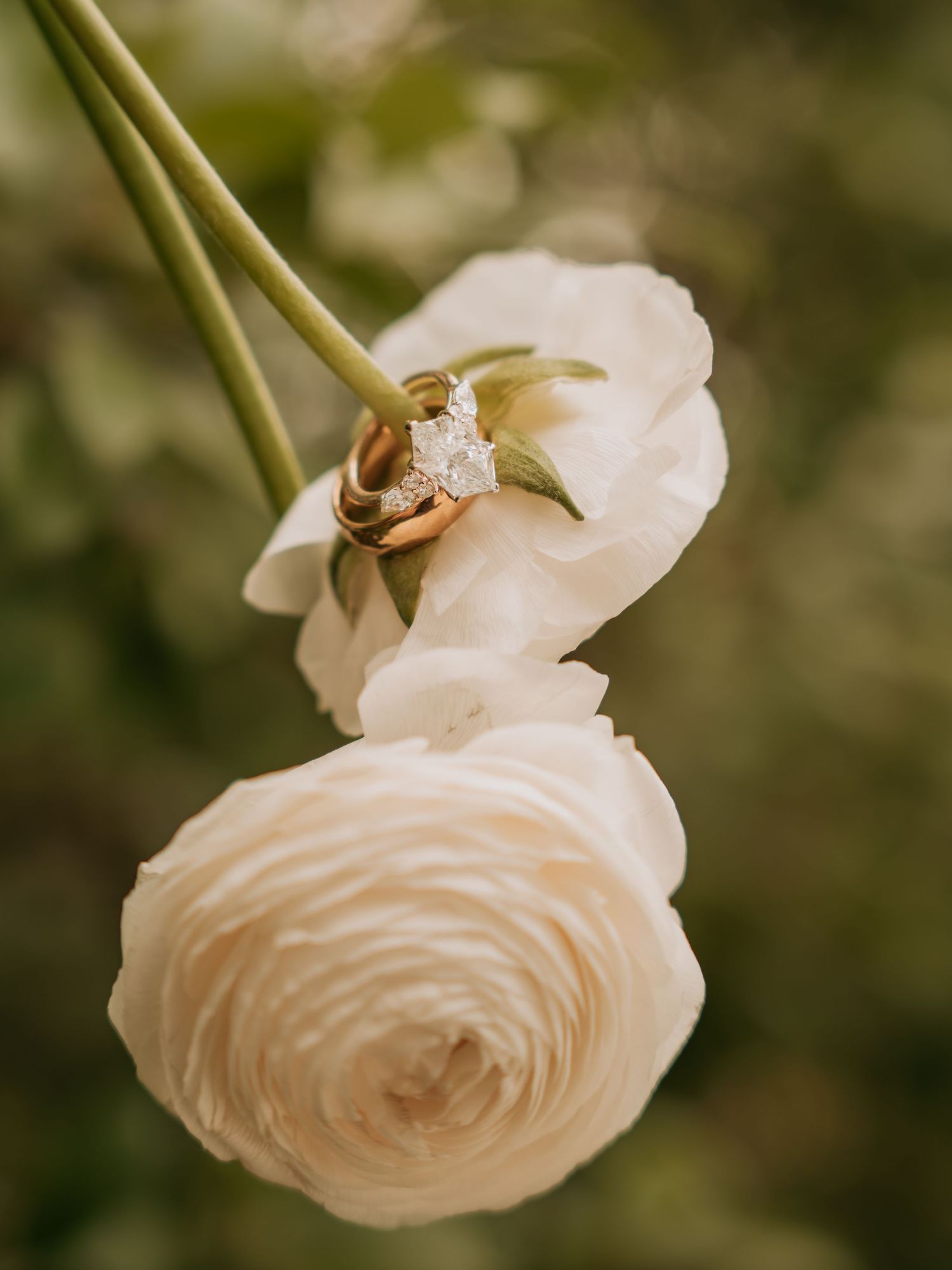 Wedding rings resting on white ranunculus flowers with a sparkling diamond engagement ring, set against a soft green bokeh background.