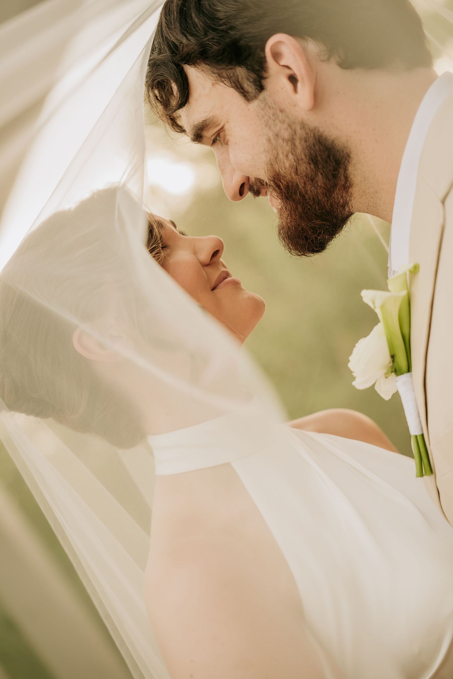 Bride and groom share an intimate moment under a flowing white veil, groom wearing a calla lily boutonniere.