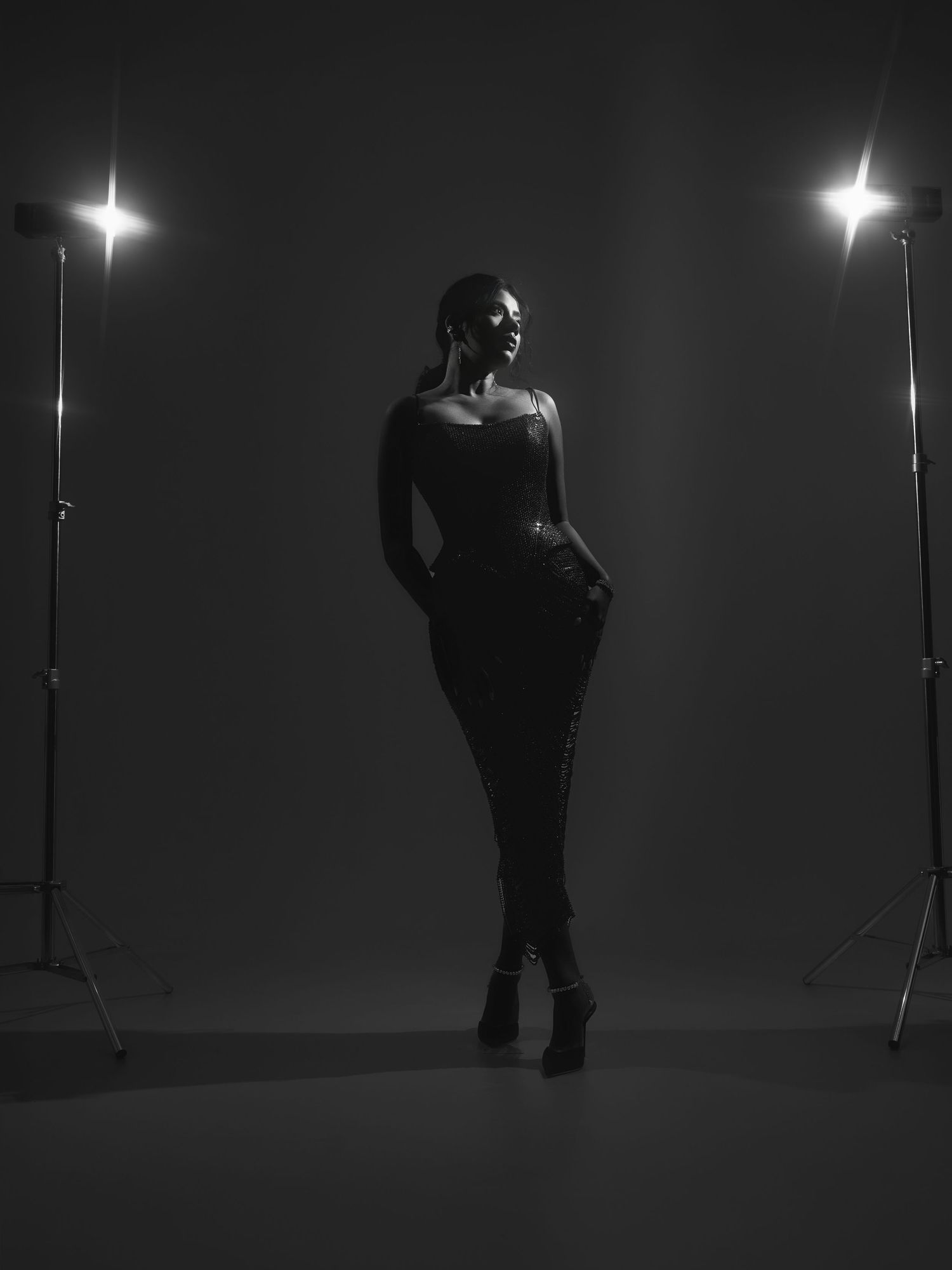 Black and white studio photo of a woman posing in dark outfit between two bright studio lights on stands.