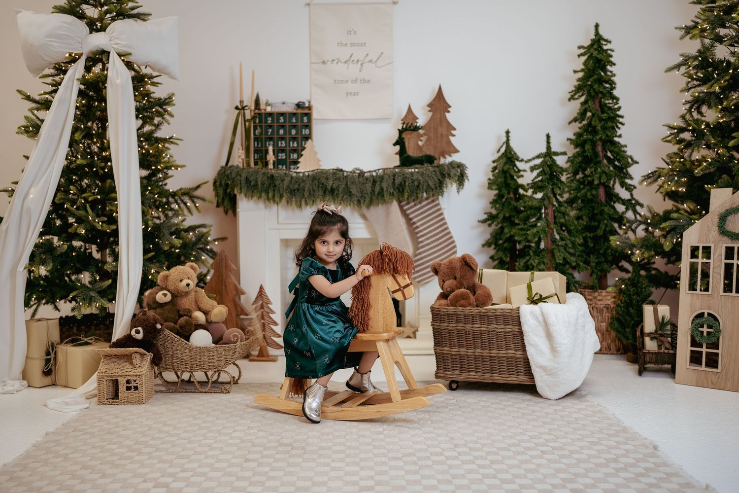 Young girl in green dress riding wooden rocking horse in festive Christmas studio with trees and teddy bears.