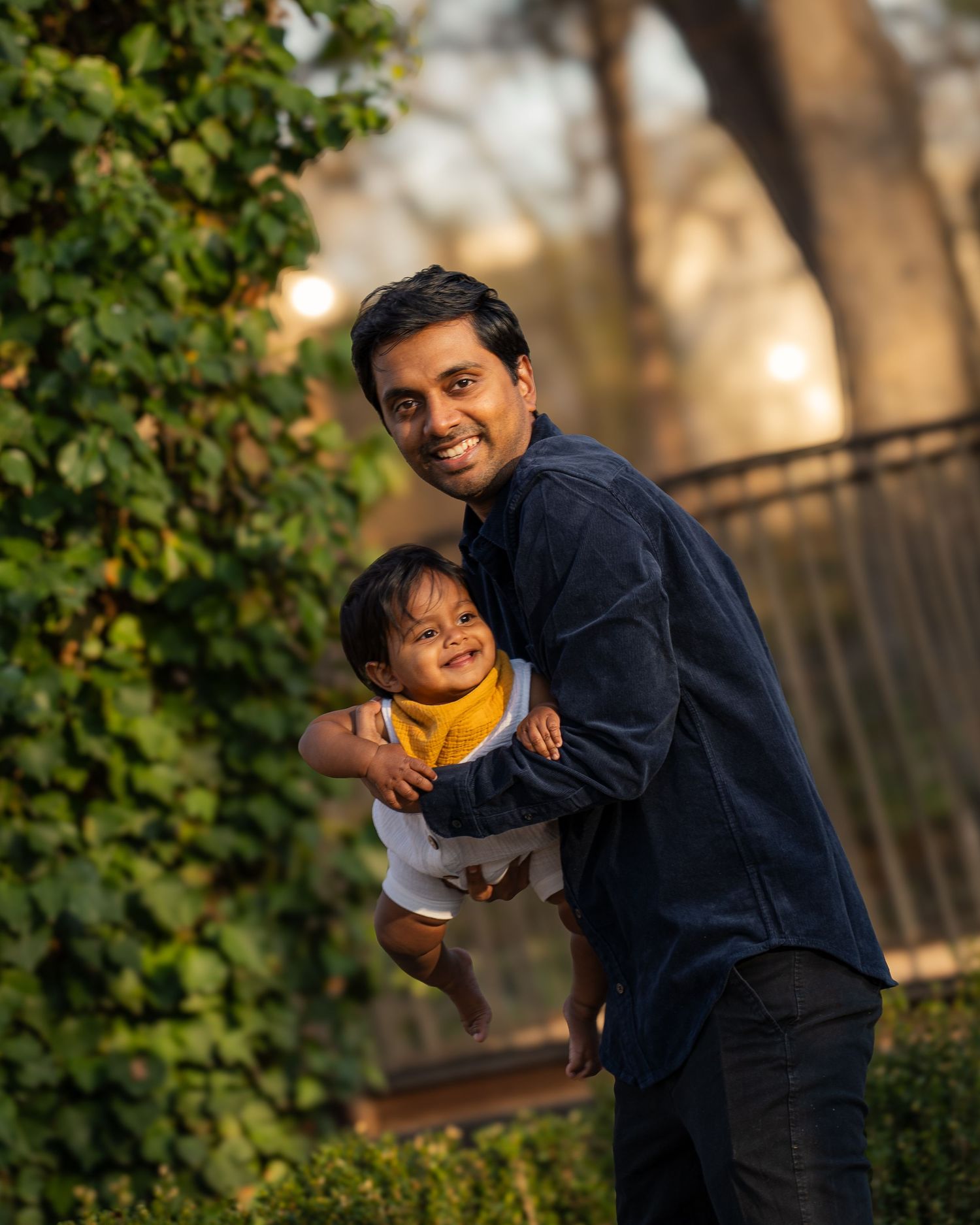 A smiling man holds a baby wearing a yellow bib outdoors, surrounded by lush green foliage in warm sunlight.
