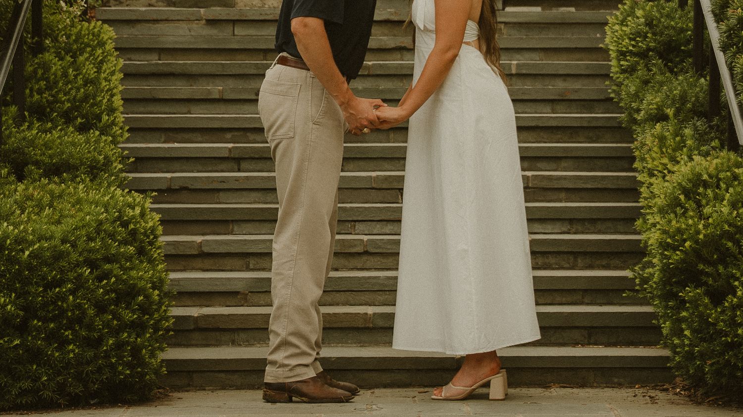 Couple holding hands in front of stone steps surrounded by lush green bushes, romantic outdoor engagement photo.