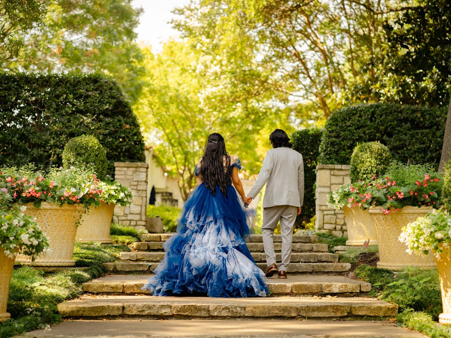 Couple walking through garden path with dramatic blue dress flowing in sunlight.