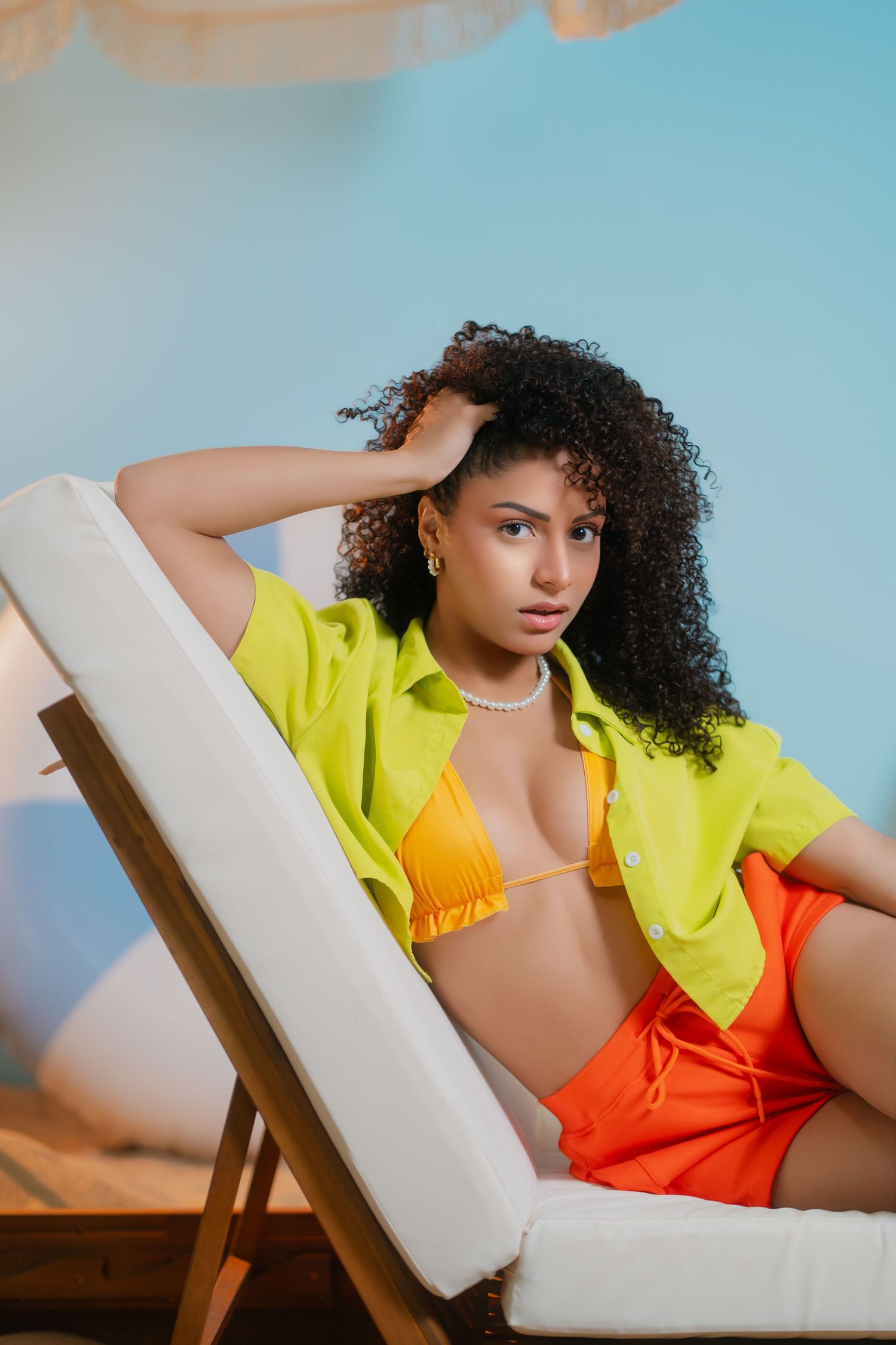 Woman in orange bikini top, yellow shirt, and orange shorts relaxing on a lounge chair against a blue background.