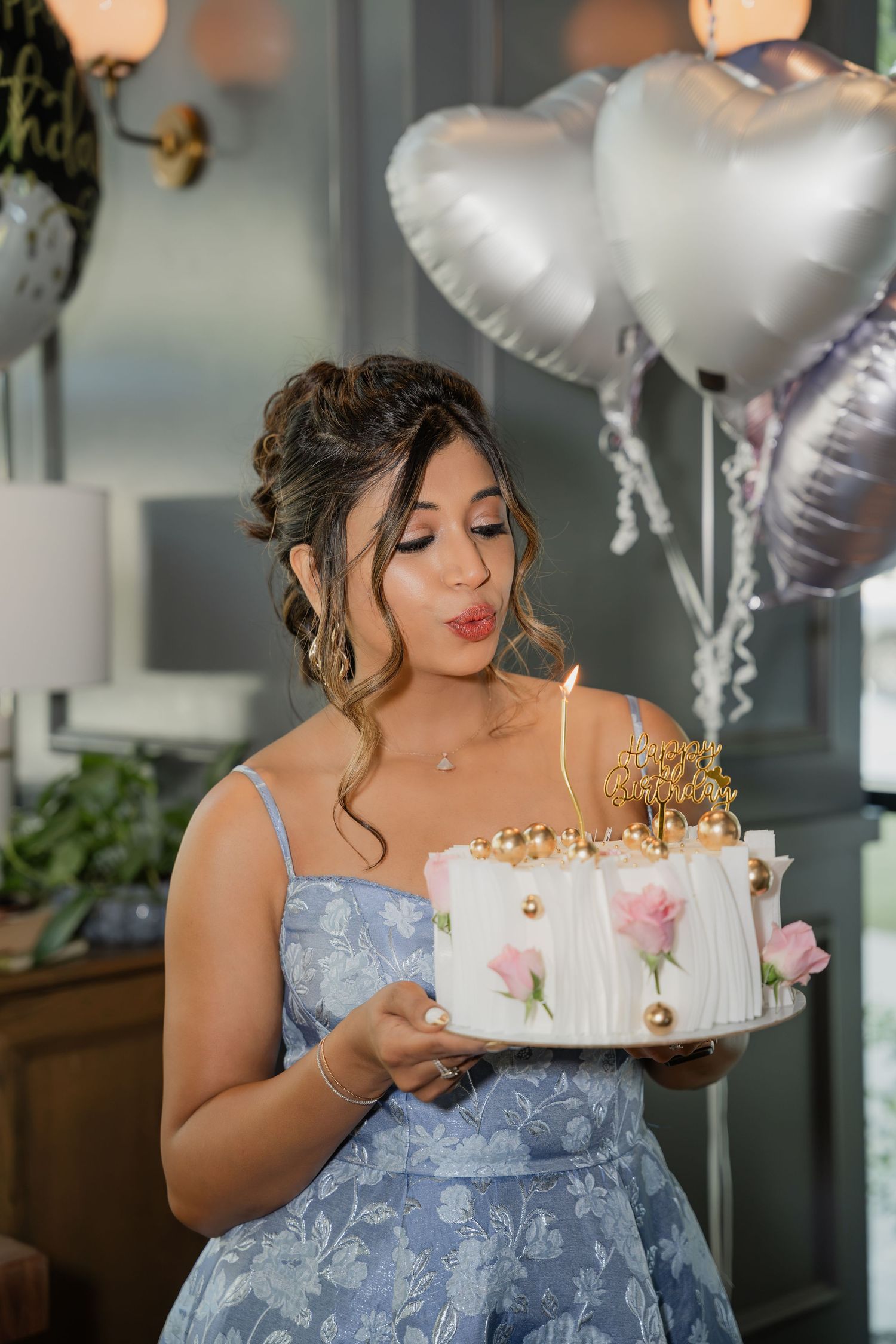 Young woman in blue floral dress blowing birthday cake candle, heart balloons in background.